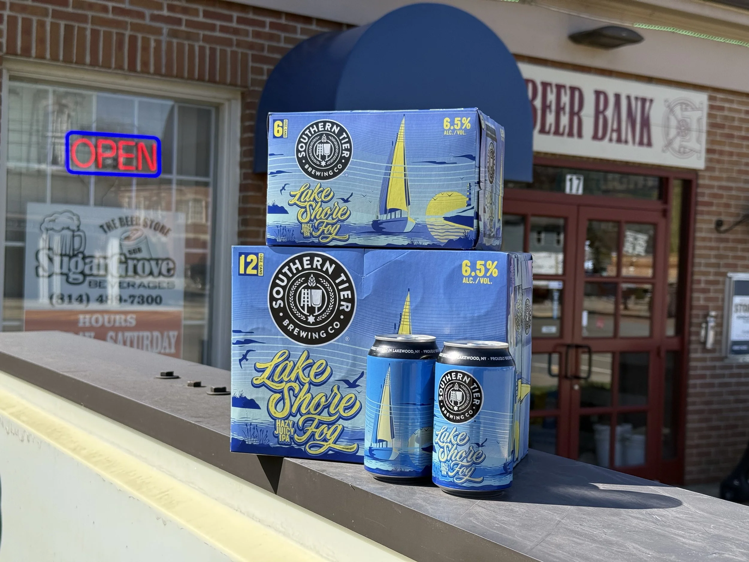 Three cans of Southern Tier Lake Shore Fog hazy juicy IPA beer stacked outdoors on a ledge, with their colorful packaging showing a sailboat and sunset. In the background, there is a brick building with a blue awning, an open sign, and a sign for a b