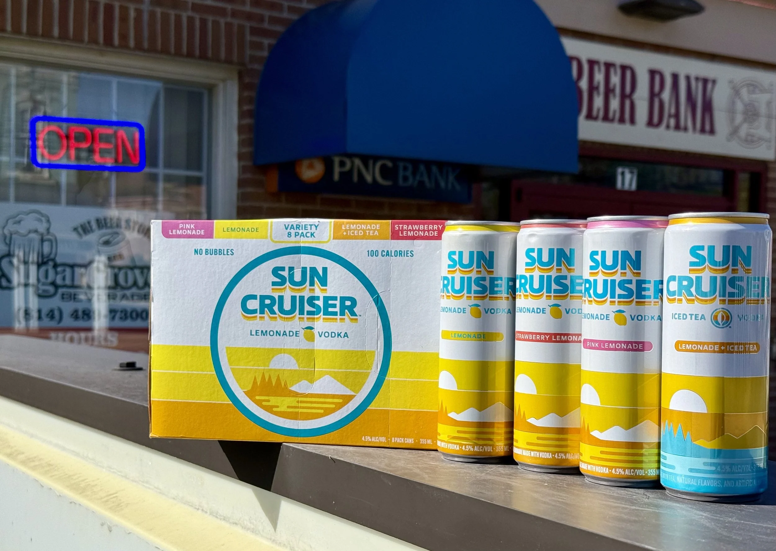 A display of Sun Cruise Lemonade cans on a store shelf with a box and signage in the background. The cans are white with colorful text and graphics, including lemon illustrations.
