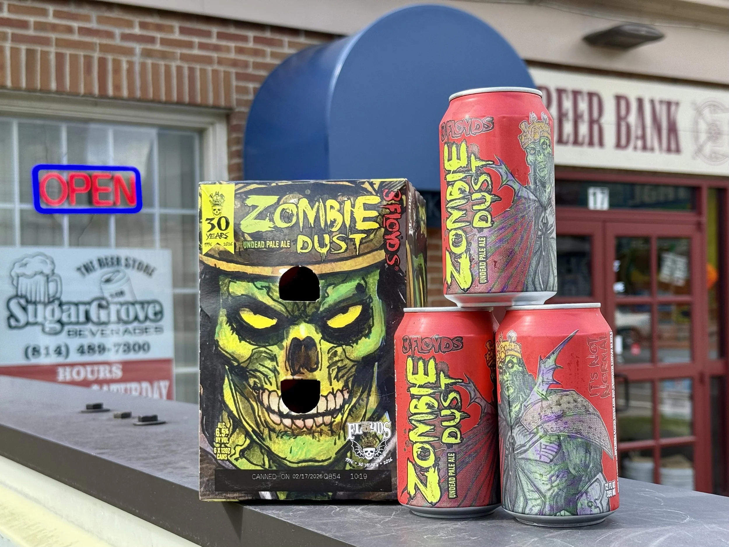 A group of Four Floyds Zombie Dust beer cans and a box with a zombie skull design displayed on an outdoor table, with a brick building, an 'OPEN' sign, and a Sugar Grove Beer and Wine store in the background.