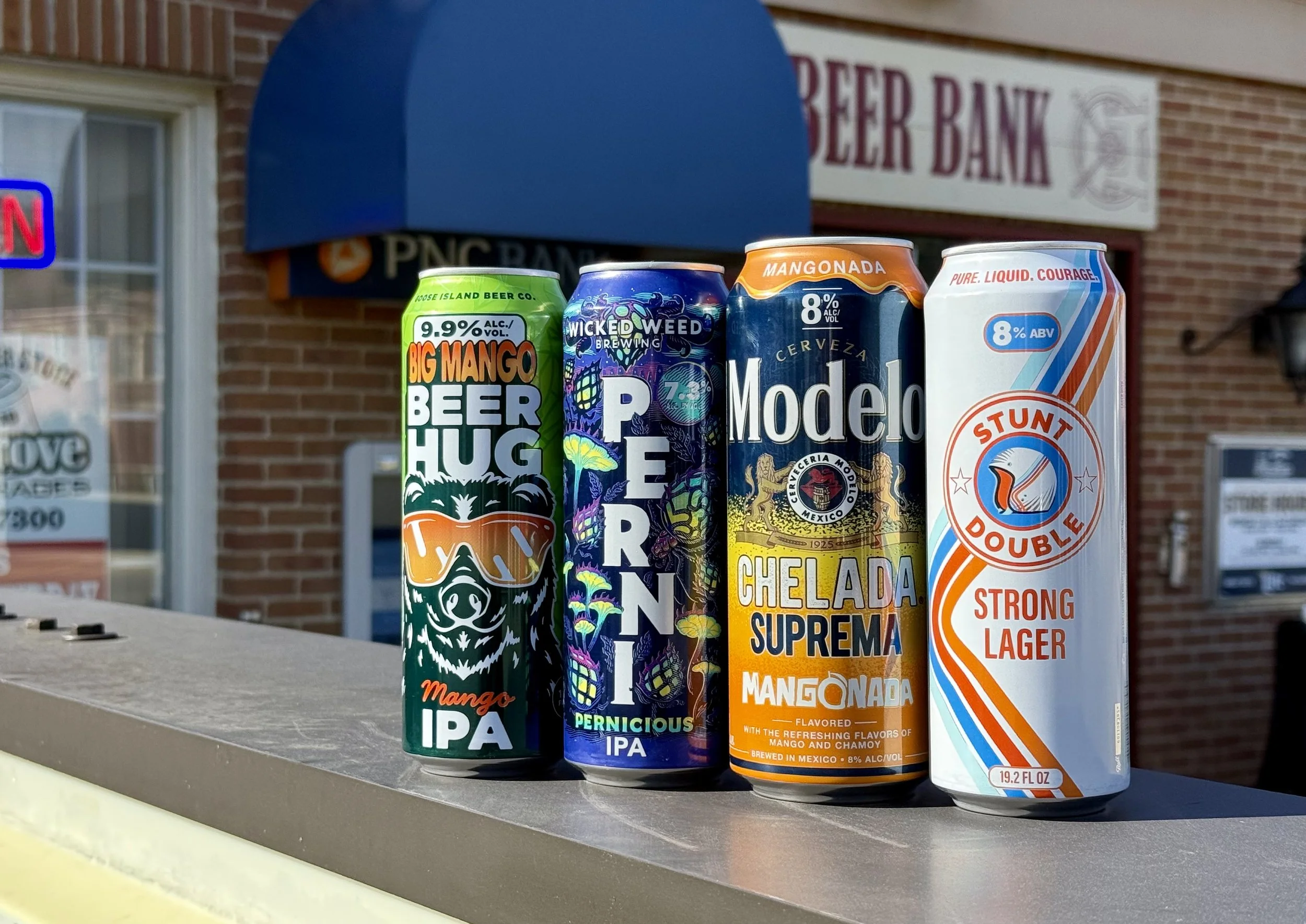 Four different beer cans displayed outdoors, with a brick building and a sign reading 'Reed Bank' in the background.