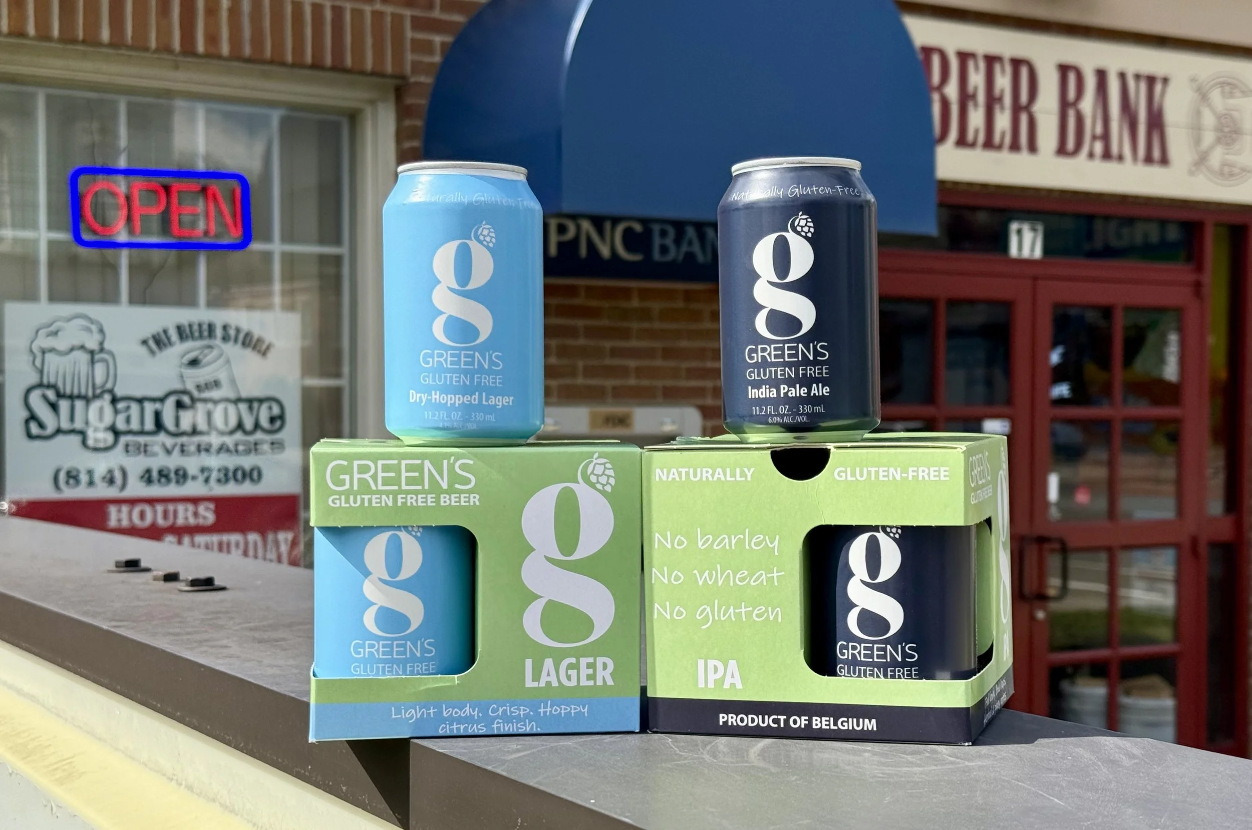 Four cans of Green's gluten-free beer, two in a light blue and two in dark blue, placed on a green box on a table outside a building with a brick wall, a window with an 'OPEN' sign, and a sign for a beer bank.