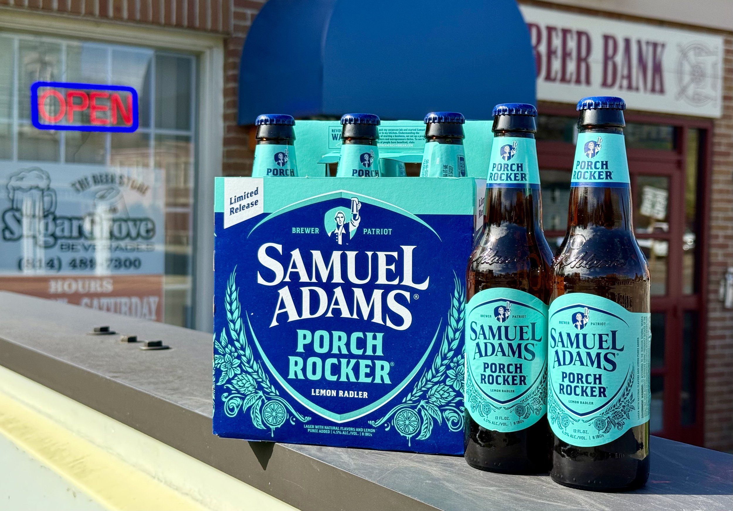 Six bottles of Samuel Adams Porch Rocker Lemon Radler beer displayed outside a store, with a blue cardboard carrier and an open sign visible in the background.