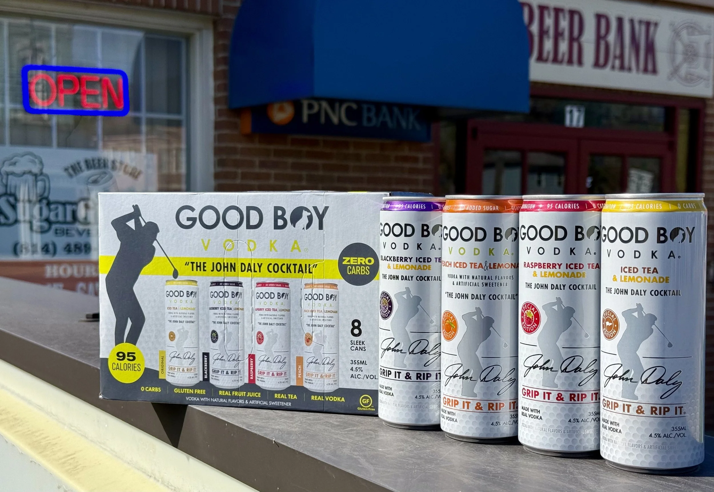 Display of canned alcoholic beverages called Good Boy Vodka, with flavors such as Blackberry Iced Tea & Lemonade, Raspberry Iced Tea & Lemonade, and Iced Tea & Lemonade, on a store shelf outside a bank.