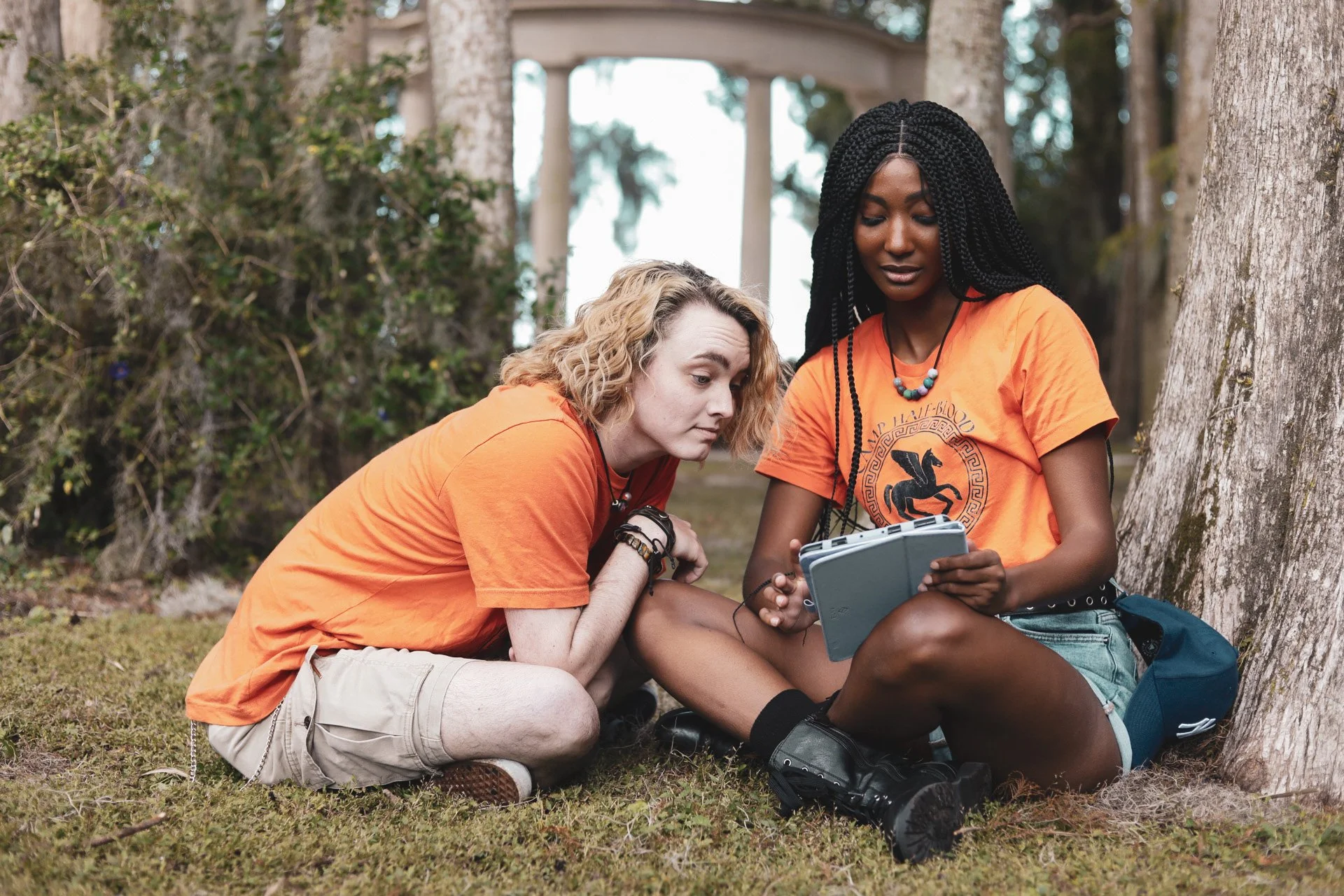 LiteraryPrincess cosplaying Annabeth Chase in box braids and an orange camp half-blood tshirt like in the pjo tv series; they are showing a tablet to a blond percy cosplayer