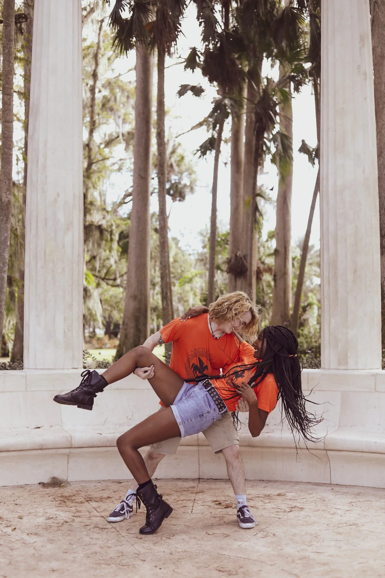 LiteraryPrincess Cosplaying Annabeth Chase in an orange camp half-blood t-shirt and black box braids with a blonde percy cosplayer dipping them and lifting their leg