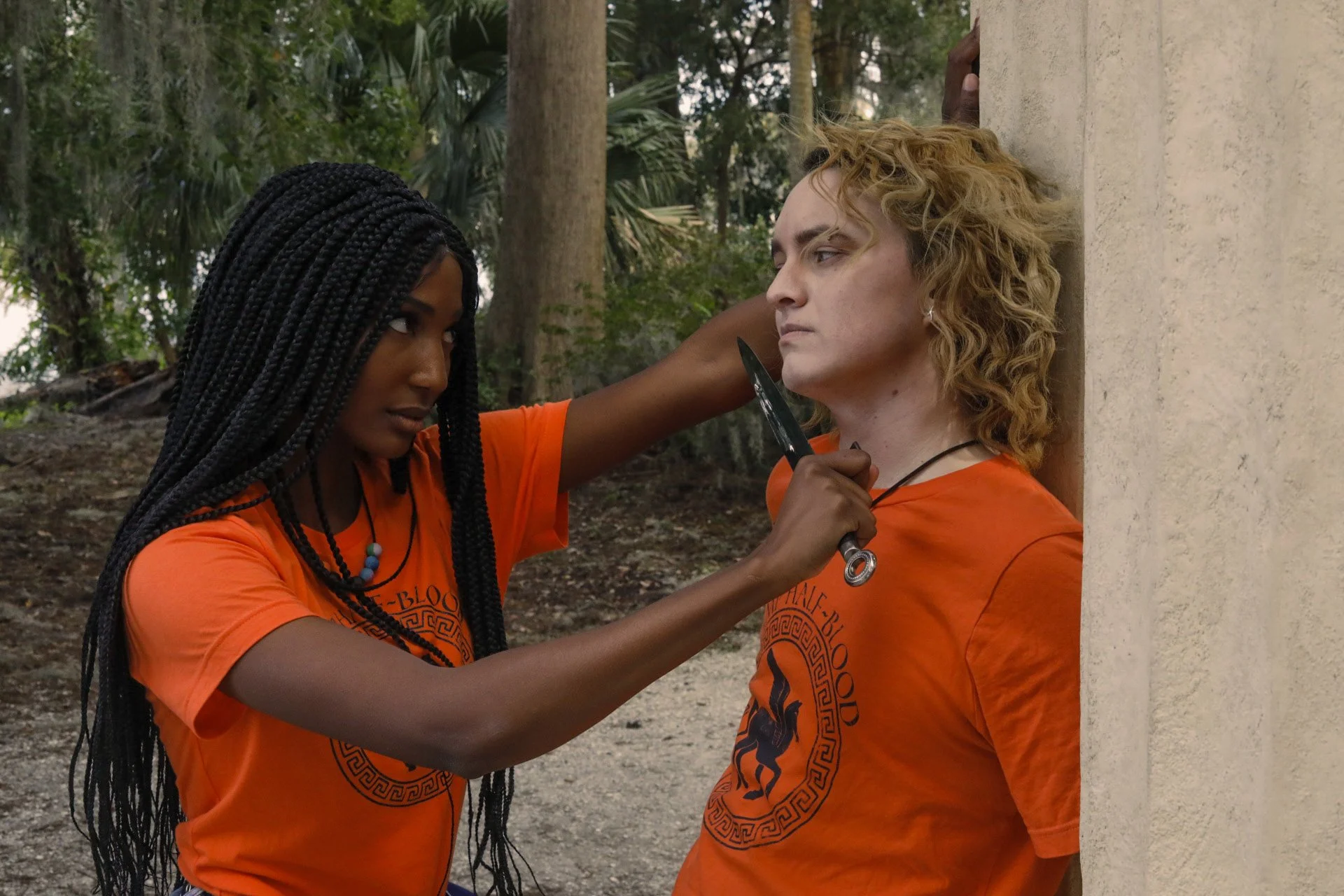 LiteraryPrincess Cosplaying Annabeth Chase in an orange camp half-blood t-shirt, similar to how she appears played by Leah Sava Jeffries in the pjo tv series. They are holding a knife to the throat of a blonde Percy cosplayer