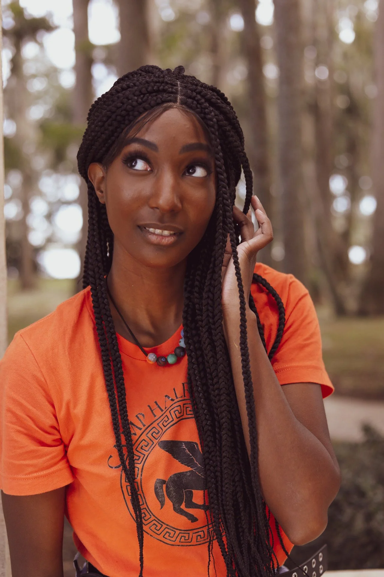 LiteraryPrincess Cosplaying Annabeth Chase in an orange camp half-blood t-shirt; they have black box braids and are tucking them behind their ear and looking up and to the side