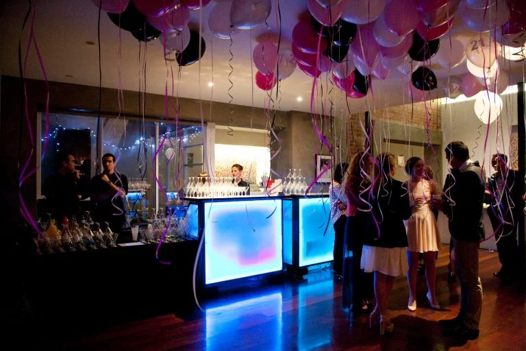 People attending a party or celebration in a decorated indoor venue with balloons, a bar, and a group of guests socializing near the bar.