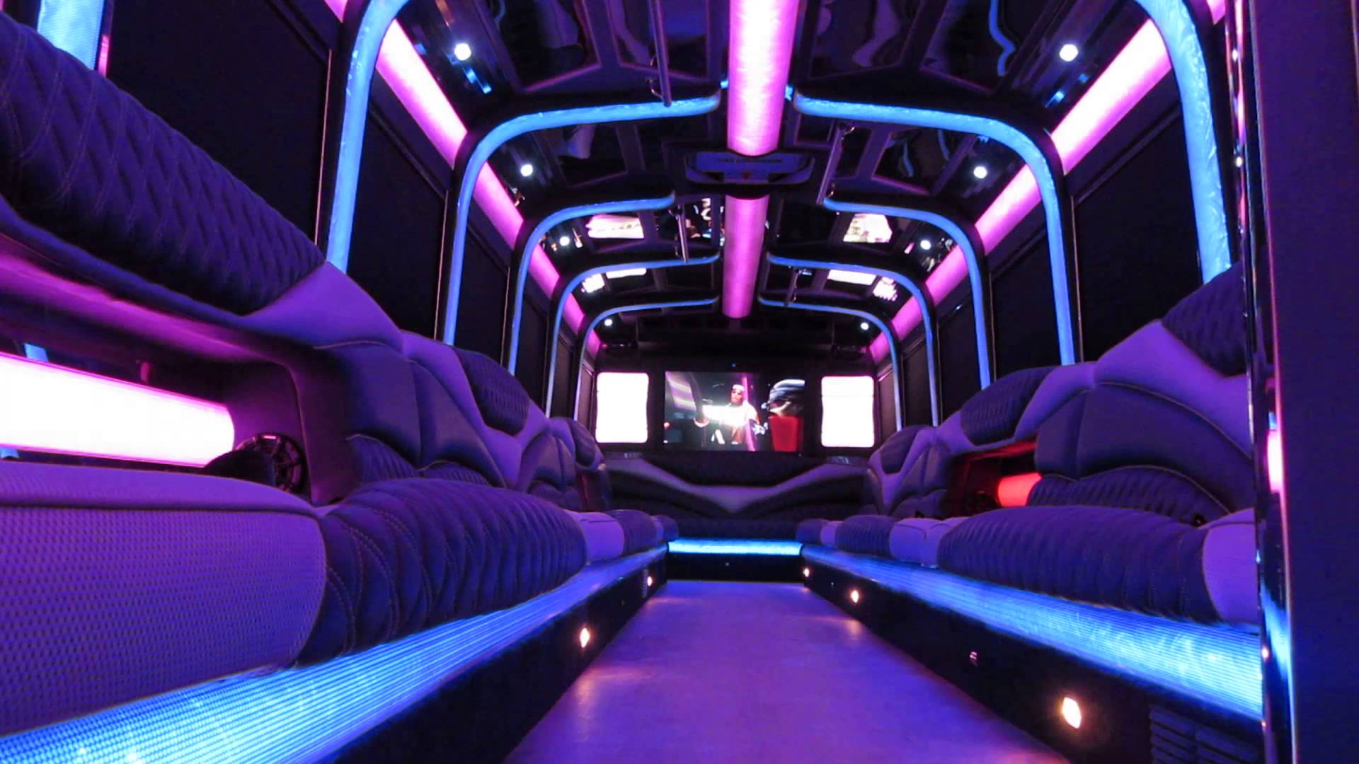Interior of a limousine bus with purple and blue neon lighting, upholstered seats, a large screen at the end, and reflective ceiling panels.