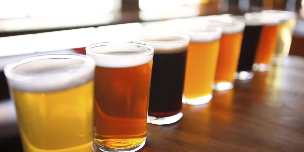 A row of seven different colored beers in glasses on a wooden table.