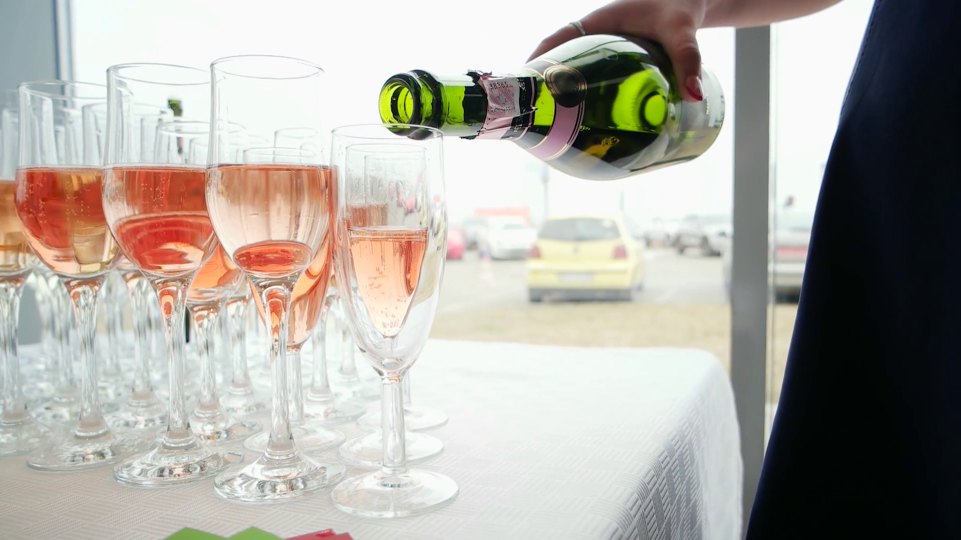 Person pouring champagne into flute glasses on a table with a white tablecloth, with a parking lot visible through the window in the background.