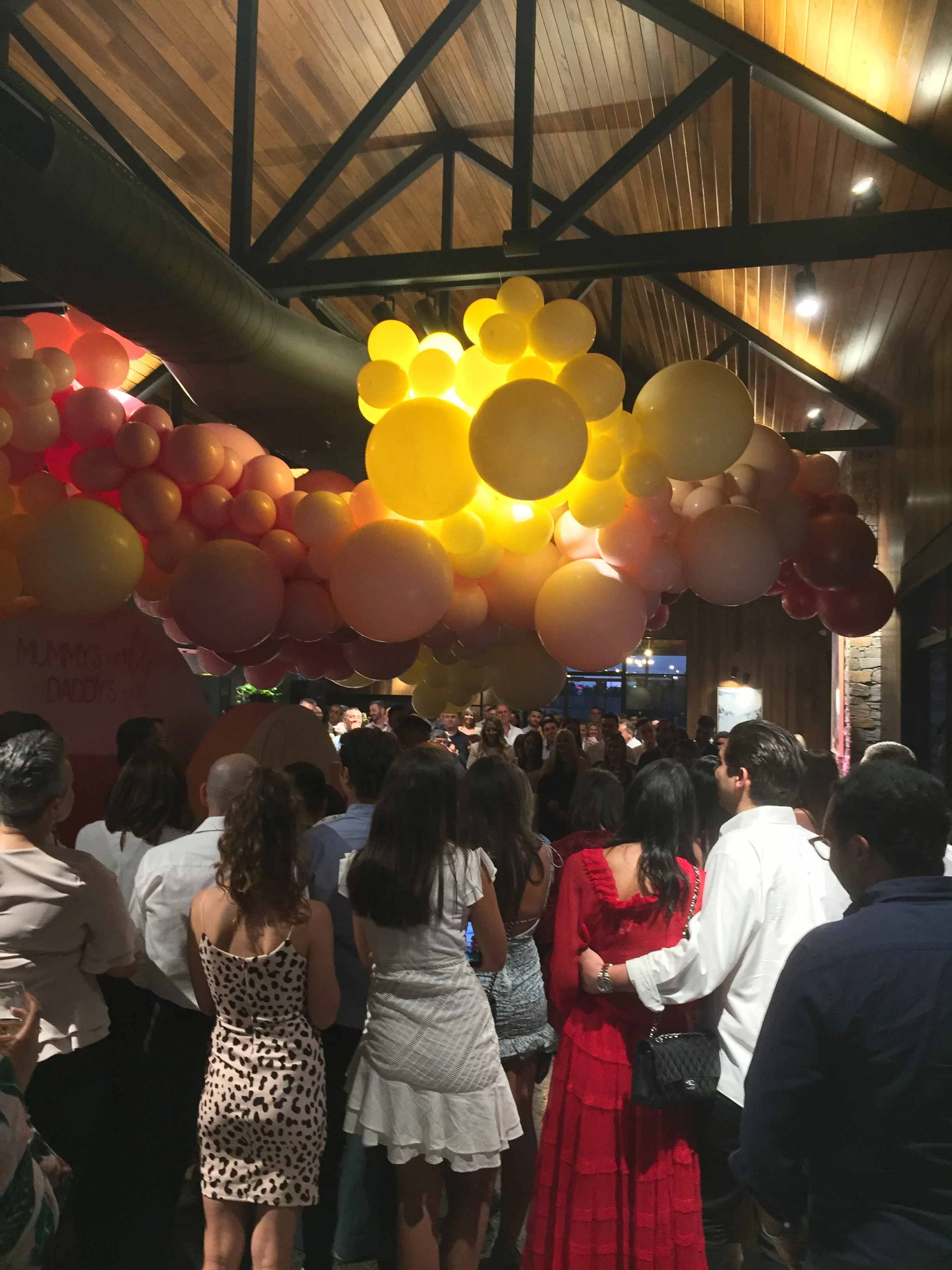 Crowd gathered at an indoor event with colorful balloon decorations hanging from the ceiling.