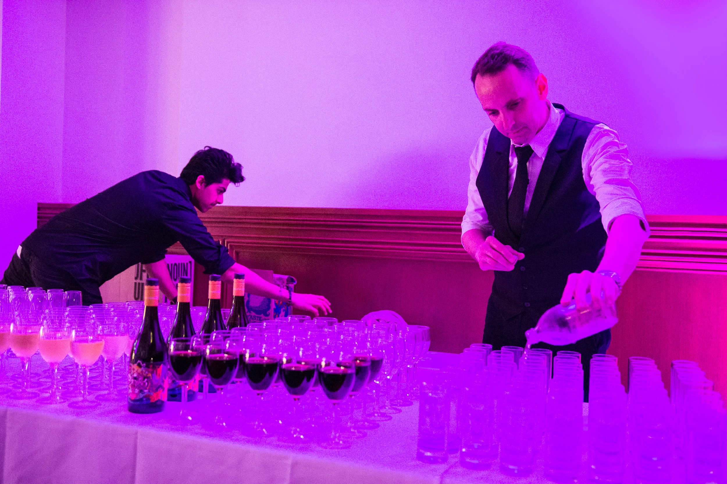 Two men in formal attire preparing drinks at a table with wine bottles and glasses, illuminated with purple lighting.