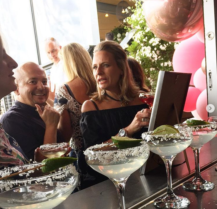 People socializing at a bar with margaritas, lime wedges, and pink balloons in the background.