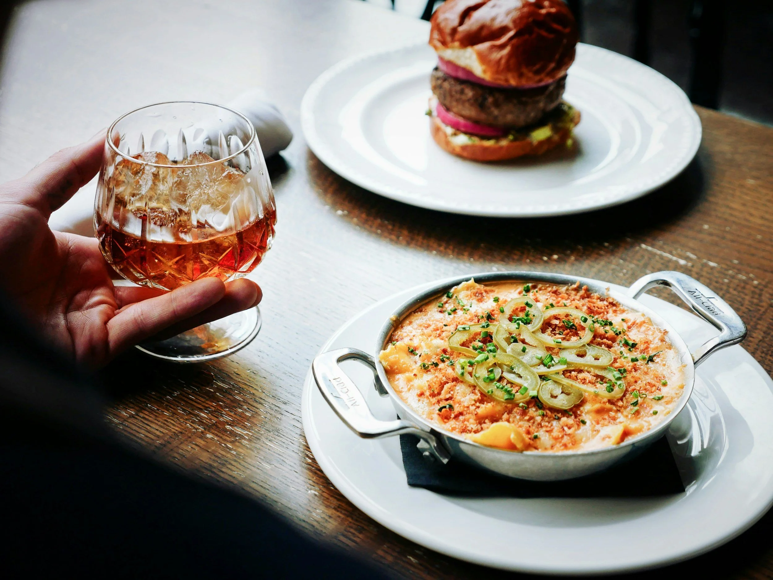 A table with a glass of whiskey, a casserole dish of baked dip topped with green onions, a cheeseburger with pickles, onions, and a beef patty on a bun.