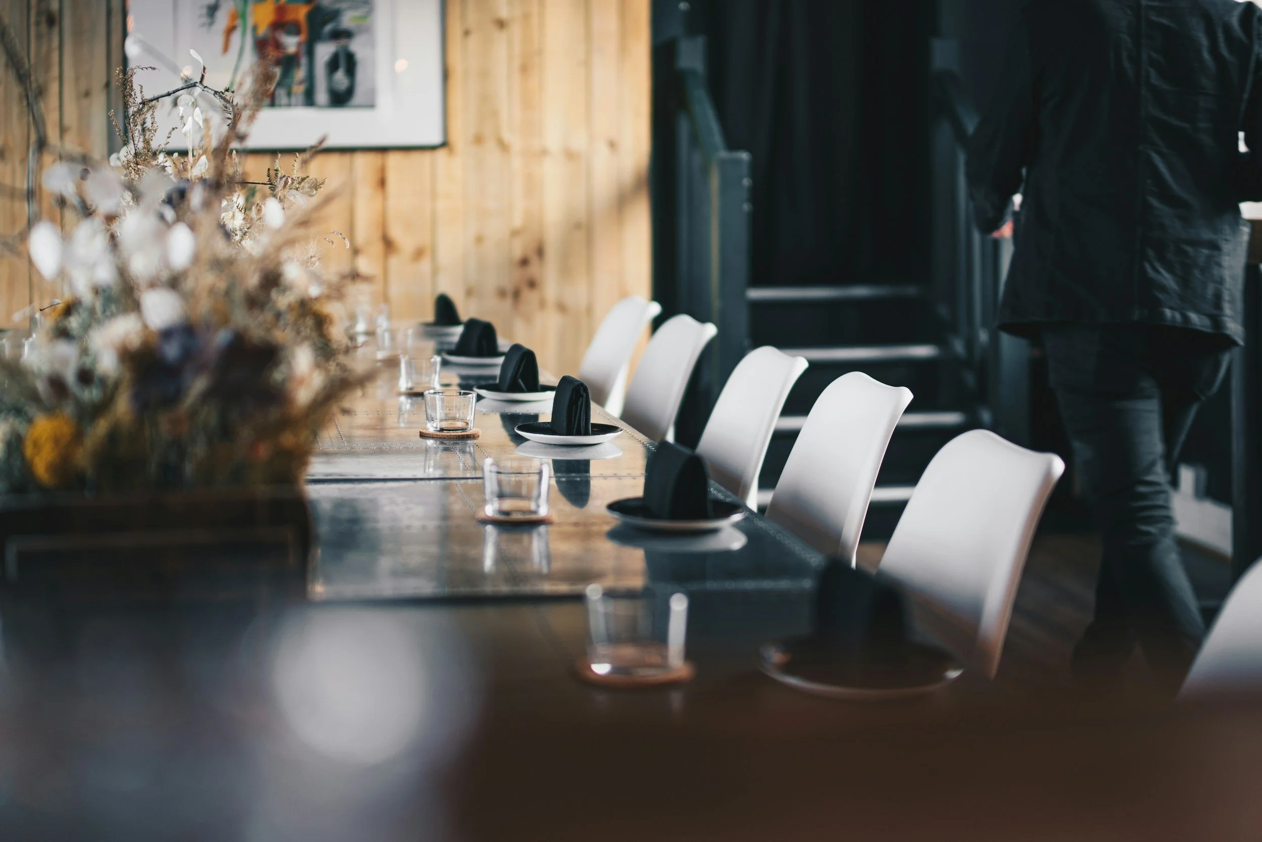 A long wooden dining table set with black napkins and glasses, surrounded by white chairs, in a rustic restaurant with wooden walls and a floral centerpiece.