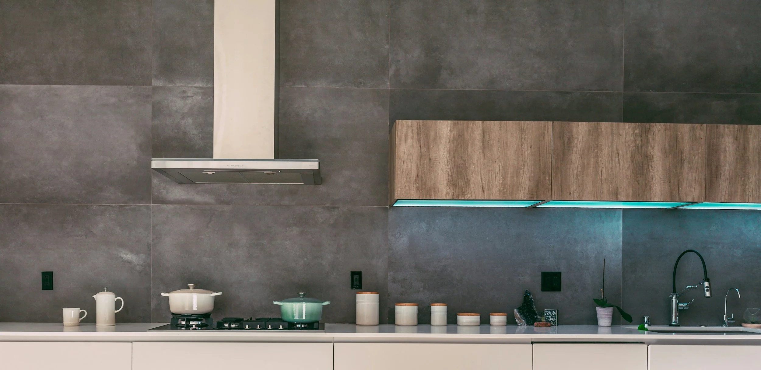 Modern kitchen counter with white cabinetry, gray backsplash, and a wooden upper cabinet. Contains pots on stove, small cups, jars, a plant, and a sink with a faucet.