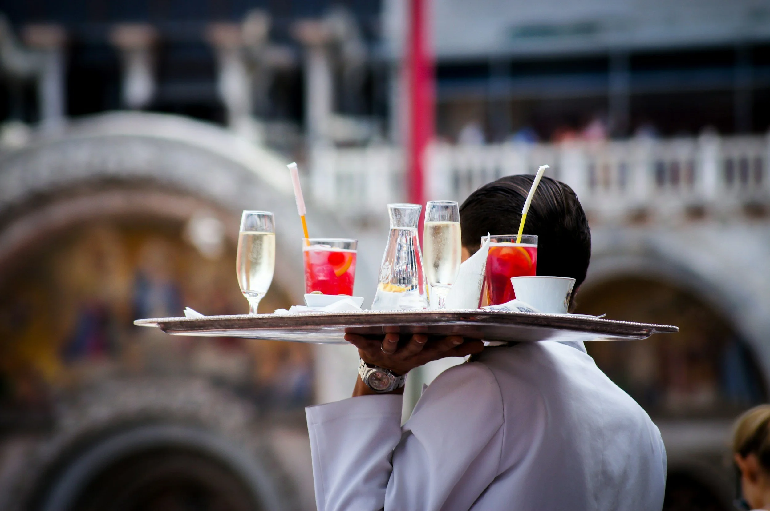 Professional waiter serving drinks on a round tray with cityscape and bridge blurred in the background, supported by Origin Five hospitality services.