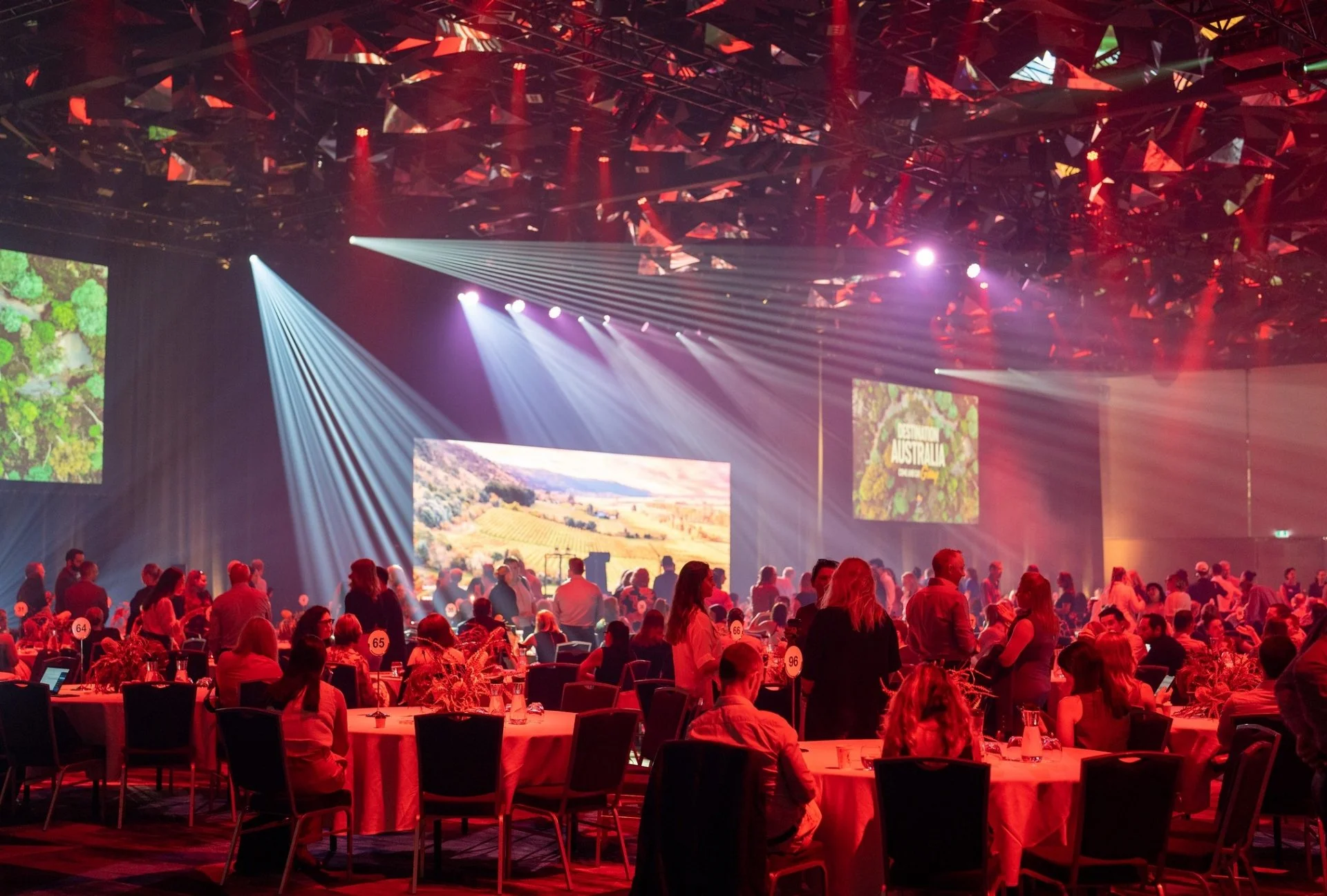 People gathered at tables with red tablecloths in a large conference hall with bright stage lights and screens displaying visuals related to Australia.