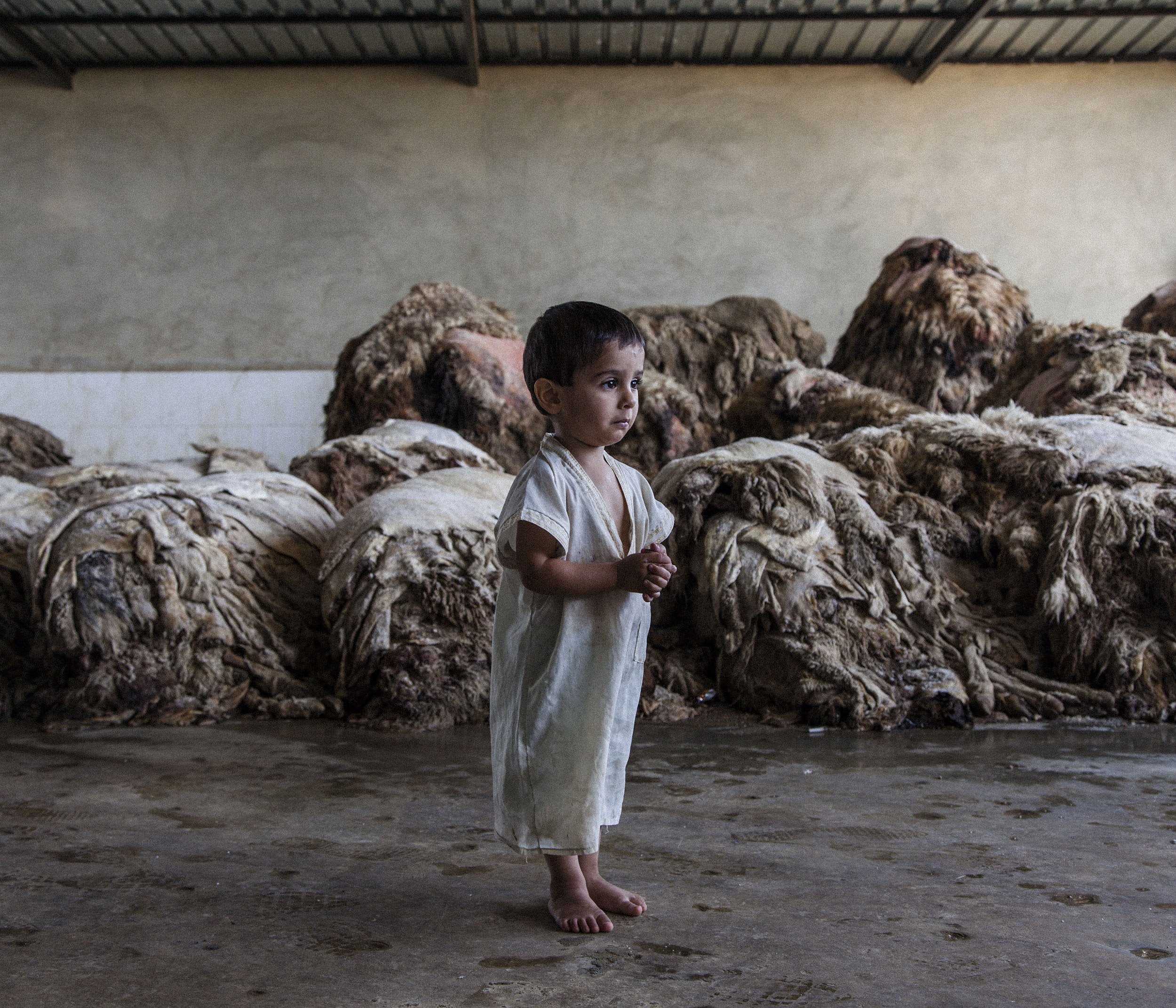  3-year-old Ali stands for a portrait in front of the piles of sheepskin which sit outside the door of his temporary home. Ali was born in the grounds of a tannery where his family live as refugees in a small two roomed apartment. Originally from Hom