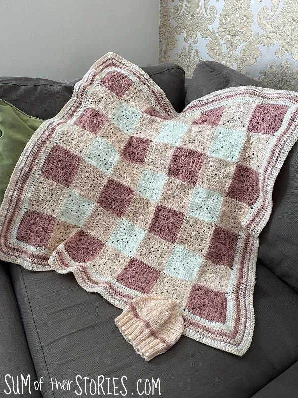 brown and cream crochet baby blanket in a gingham design laid over the back of a sofa, a little baby hat of the same yarn is in the forground
