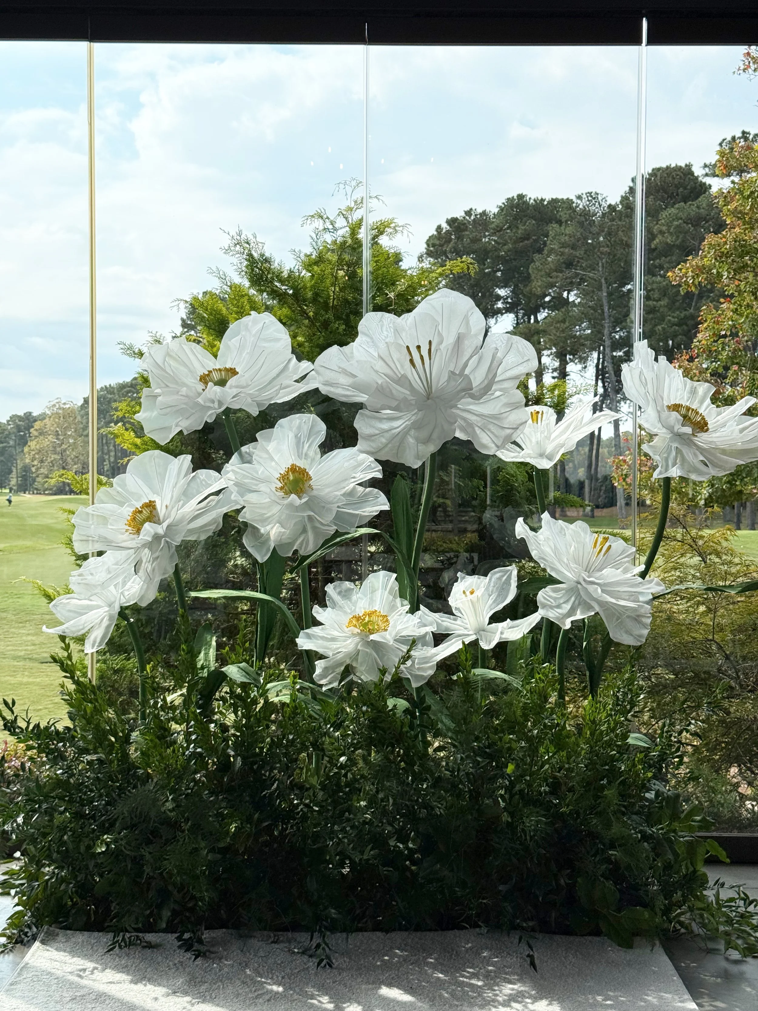 White decorative flowers displayed in front of a large glass window with a green outdoor landscape in the background.