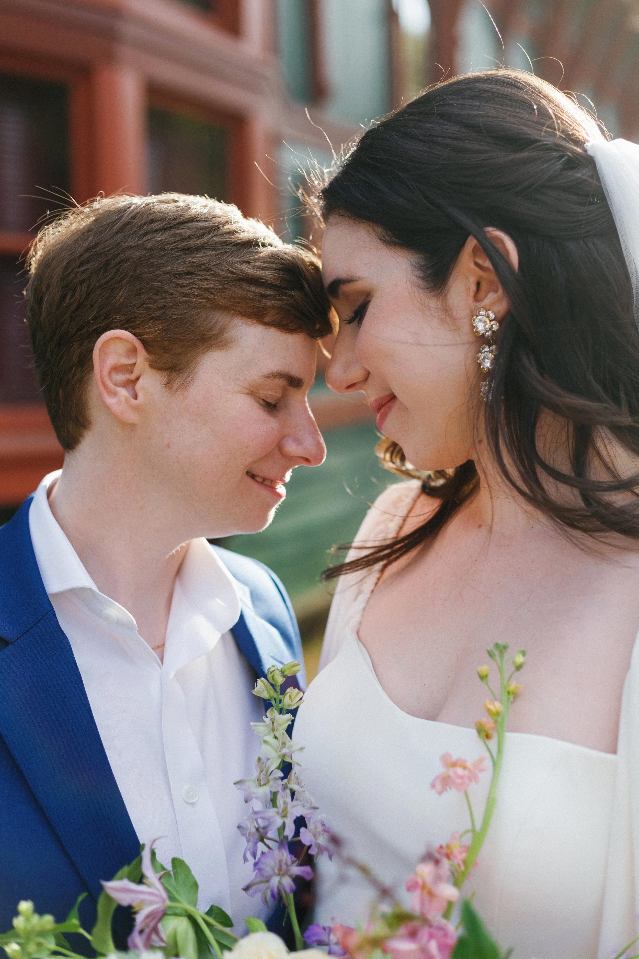A newlywed couple smiling with foreheads touching outdoors during sunset, with flowers and a rustic building in the background.