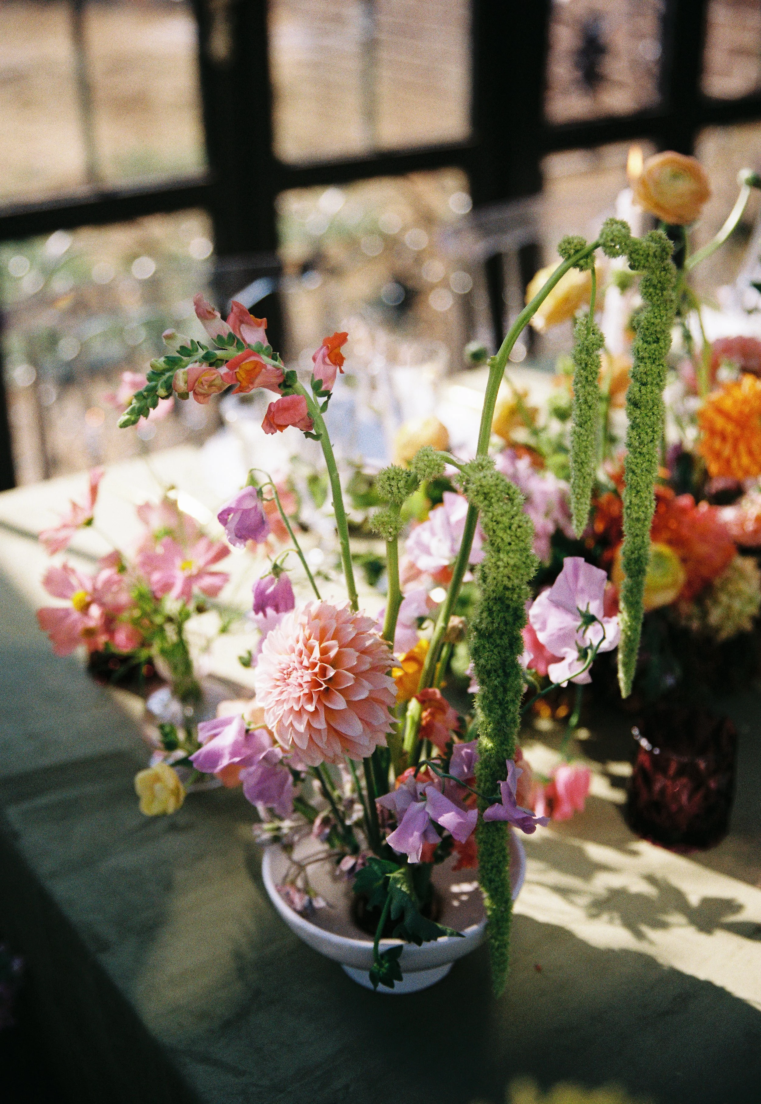 A colorful bouquet of various flowers in a white vase, placed on a table with daylight coming through a window in the background.