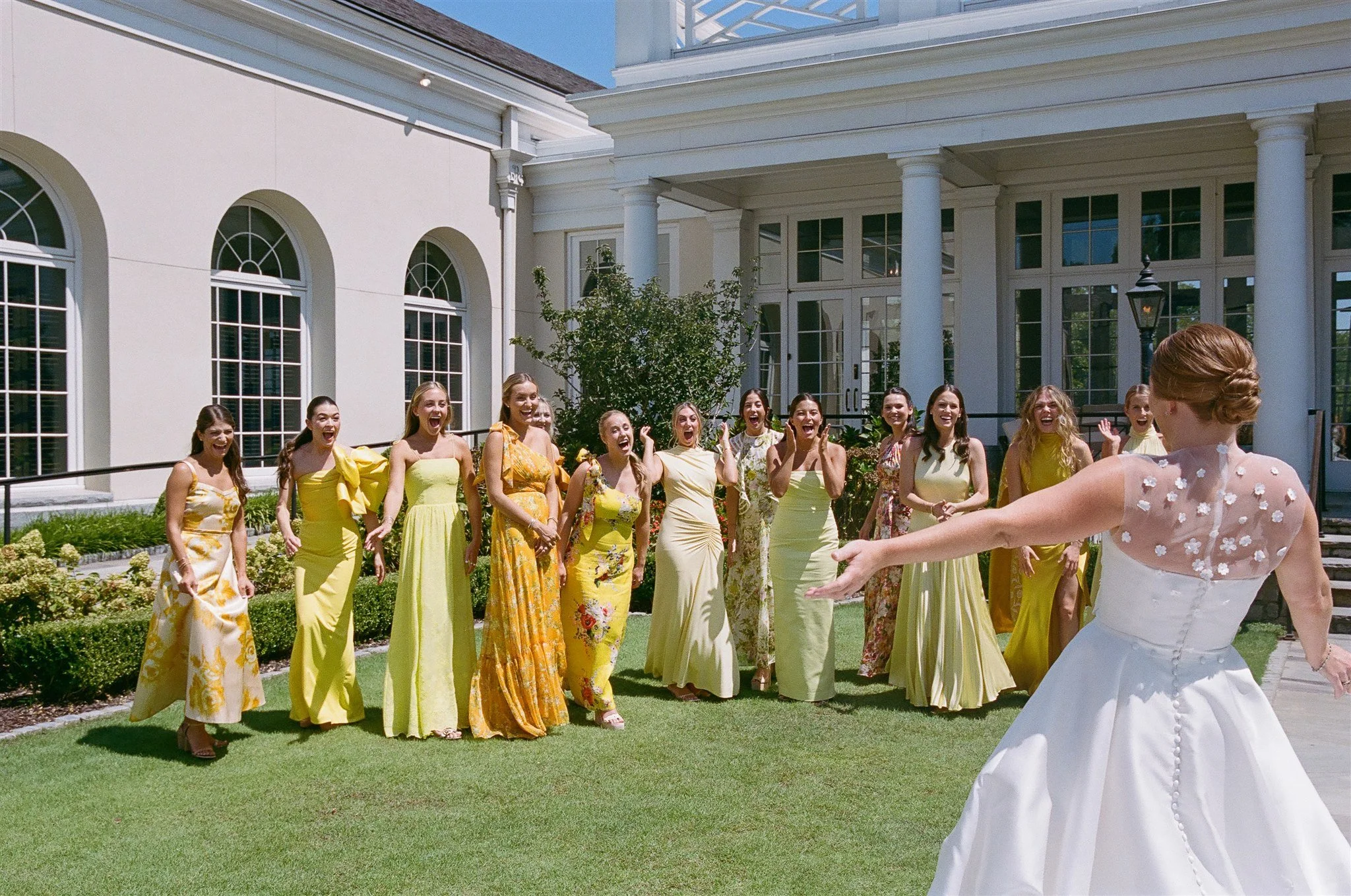 A bride in a white wedding dress is throwing her bouquet to a group of women in yellow dresses outside of a large white house on a sunny day.