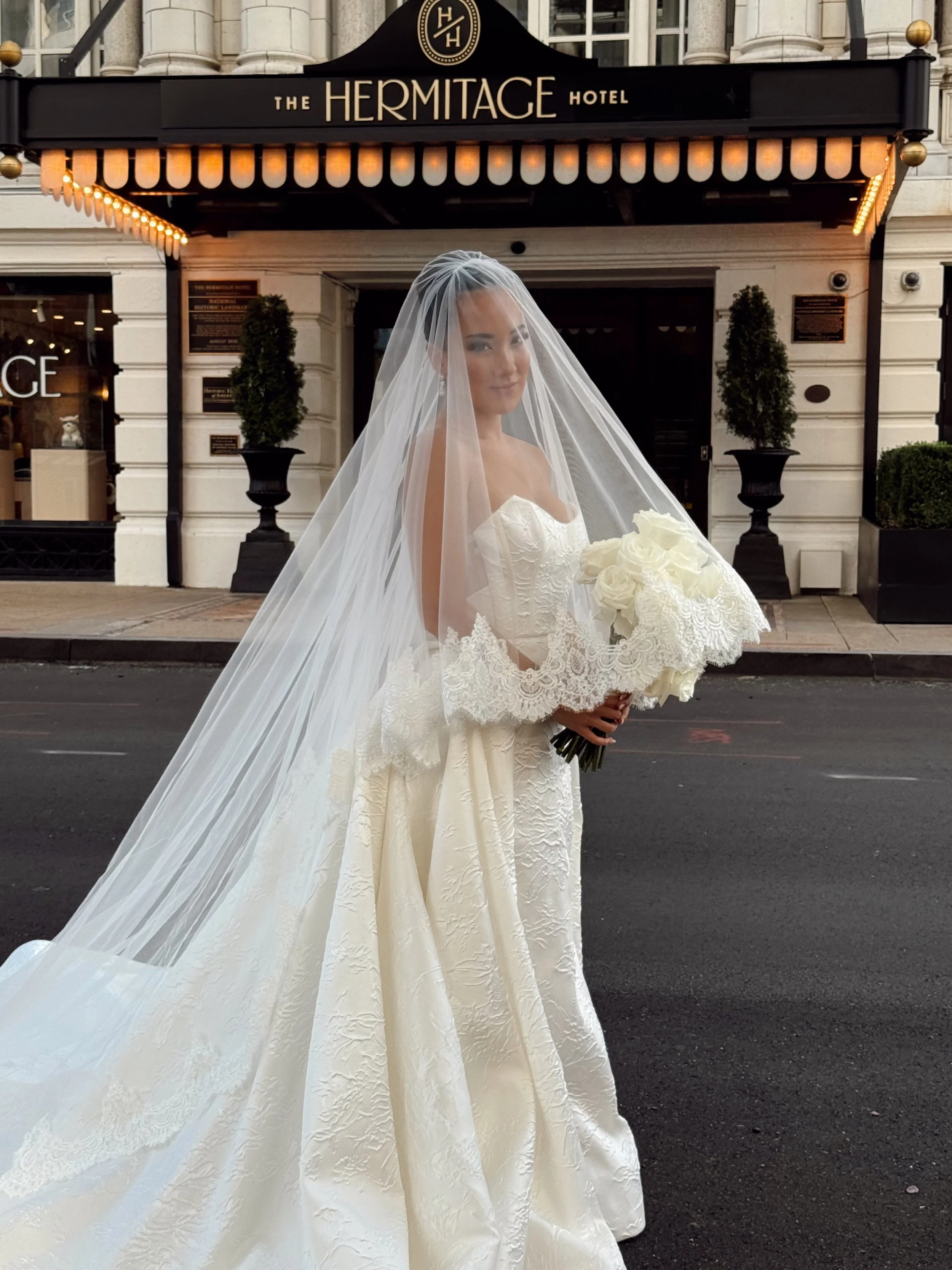 Bride in a white wedding gown with lace details, holding a bouquet of white roses, standing outside The Hermitage Hotel, with a black sign above and potted plants on either side.
