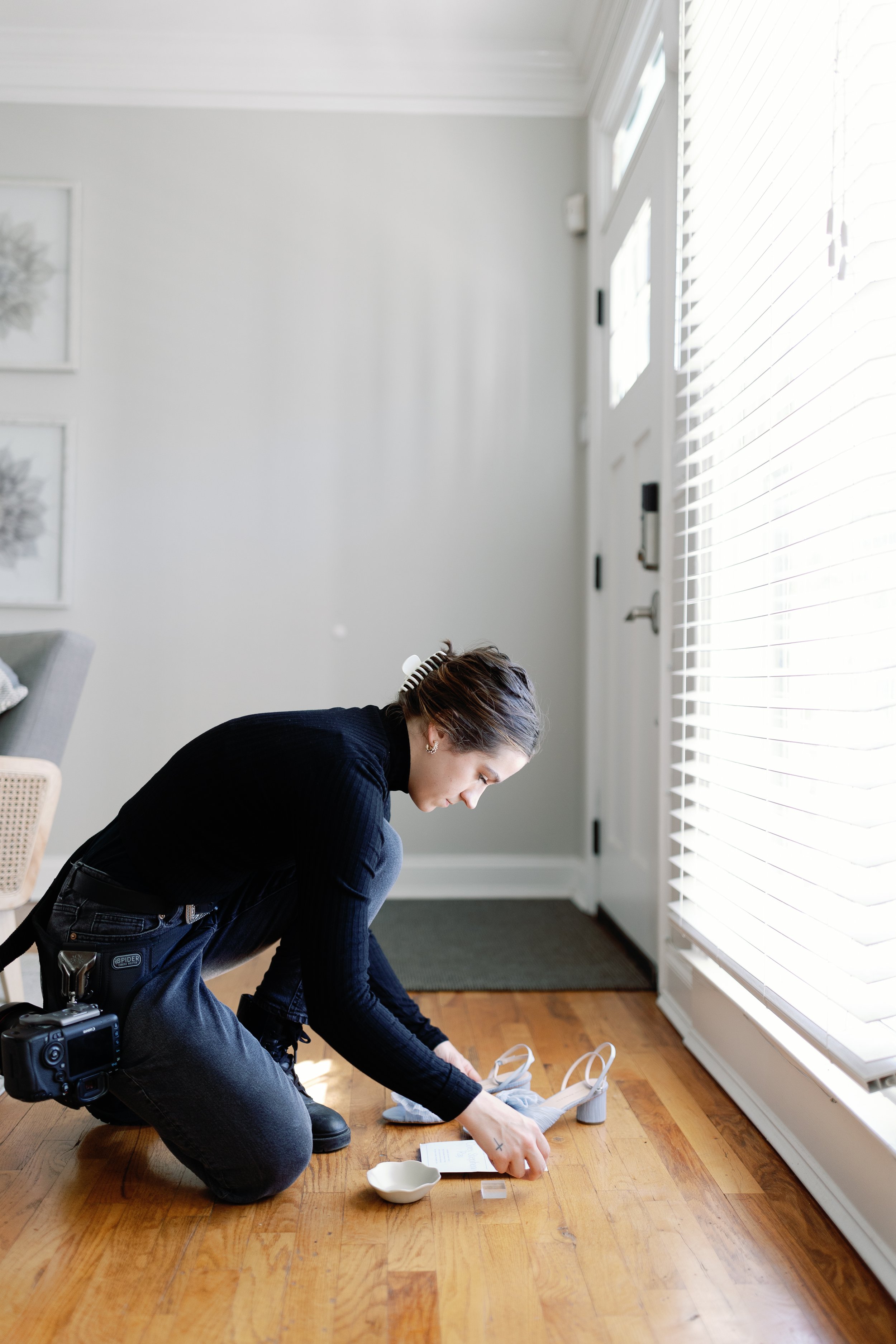 A woman kneeling on a wooden floor, bending over to adjust her light gray high-heeled shoes near a door with white blinds, with a small white dish and paper nearby.