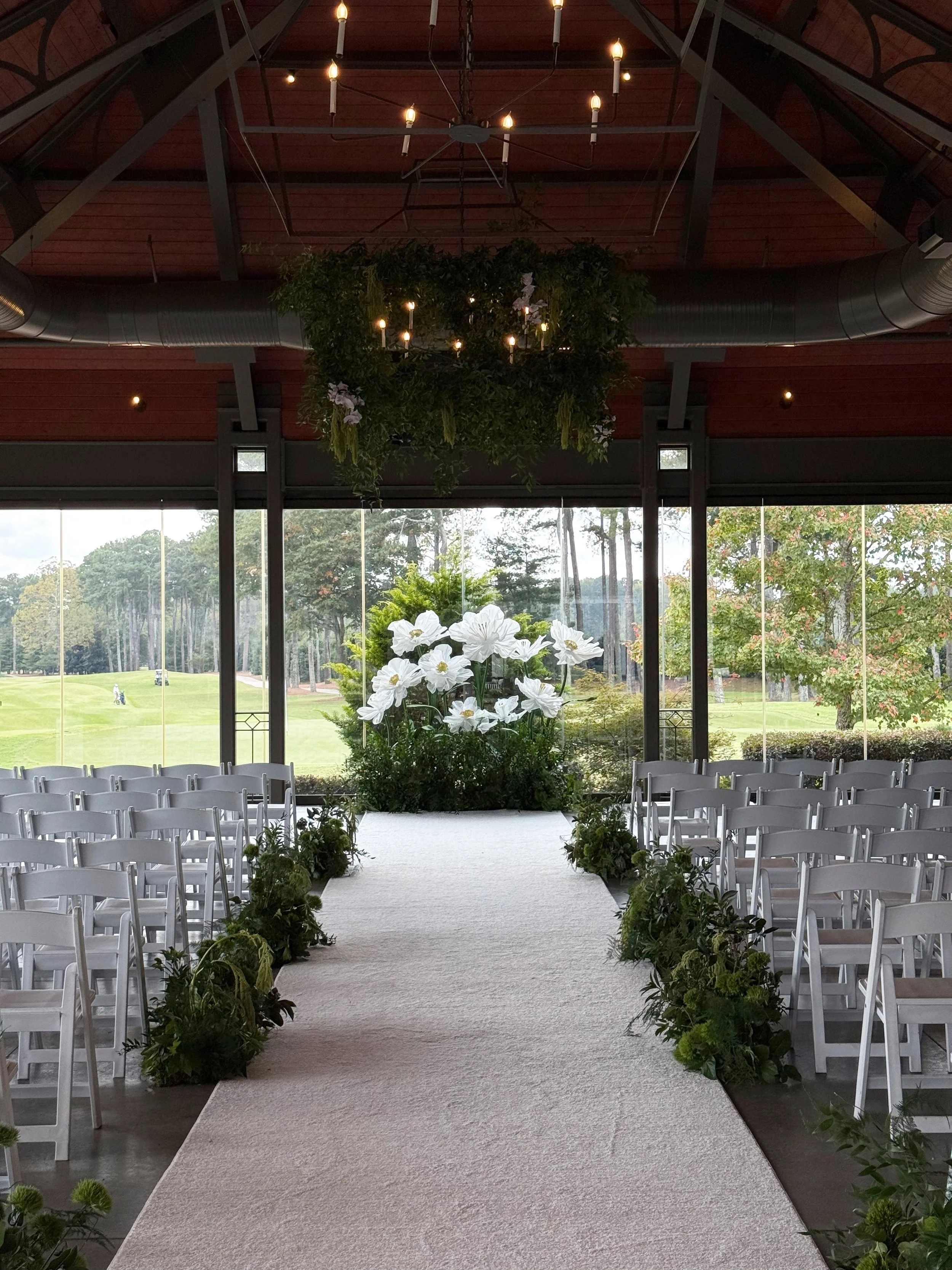 Indoor wedding ceremony setup with white chairs arranged on either side of a white aisle, decorated with greenery. A large floral arrangement of white flowers is at the altar, with a large chandelier hanging from the ceiling and large windows showing a view of a golf course or park.