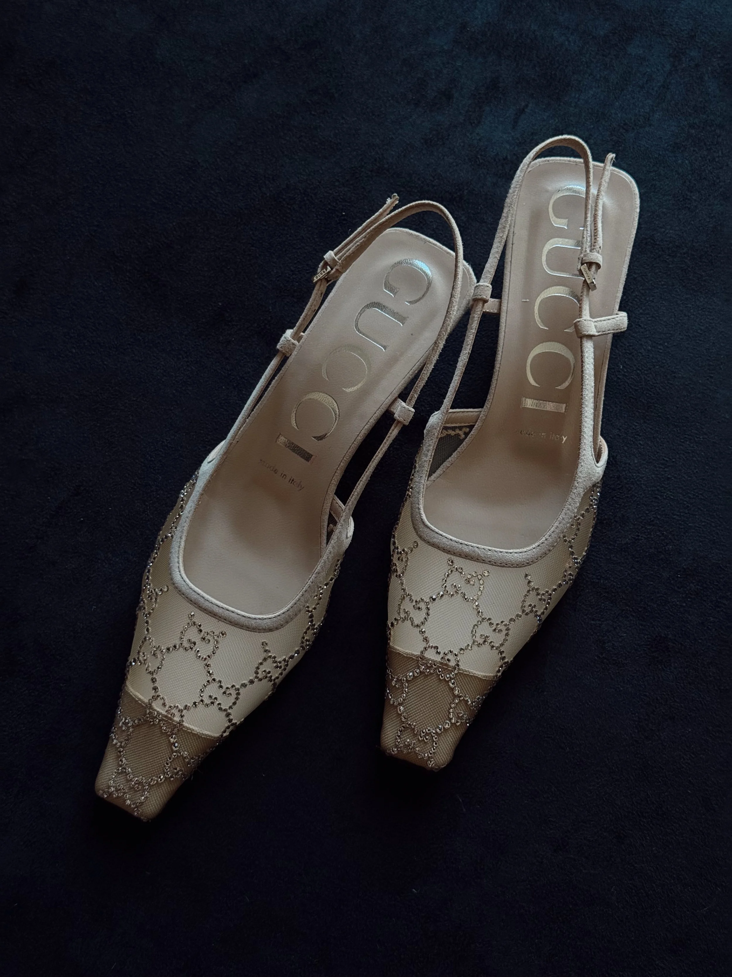 Pair of beige Gucci high-heeled shoes with pointed toes, decorative chain patterns, and ankle straps on a dark surface.