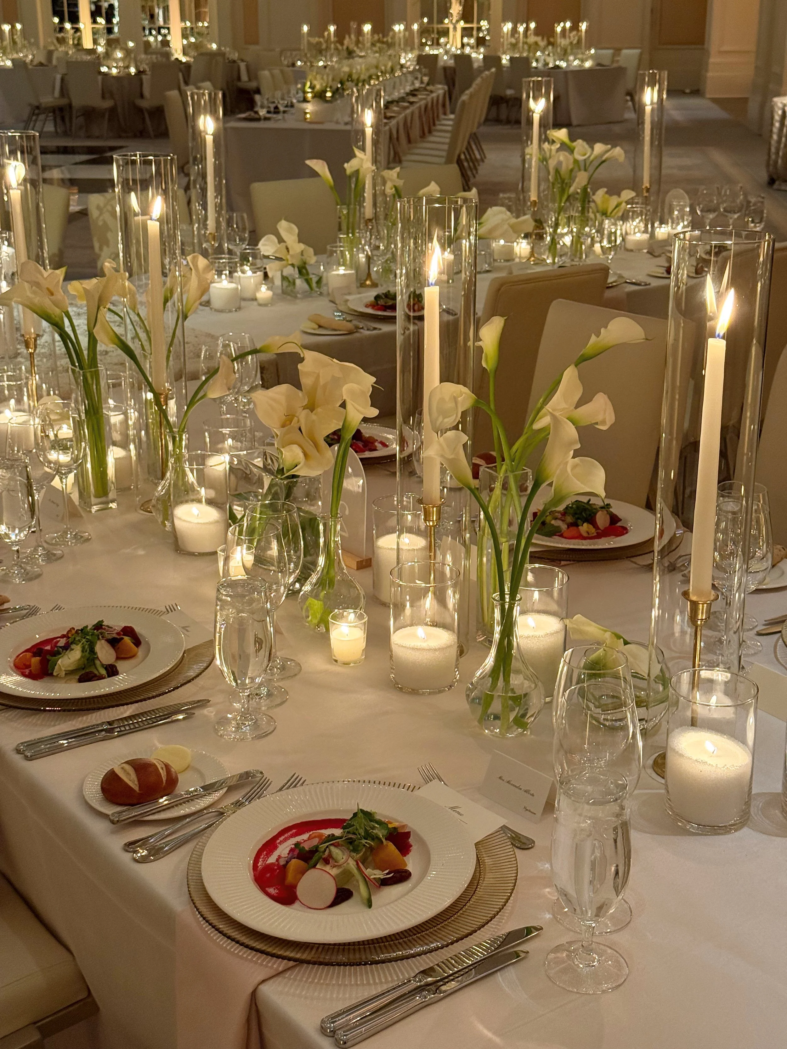 Elegant banquet table decorated with white flowers, tall candles, and glowing candles in glass holders in a formal event setting.