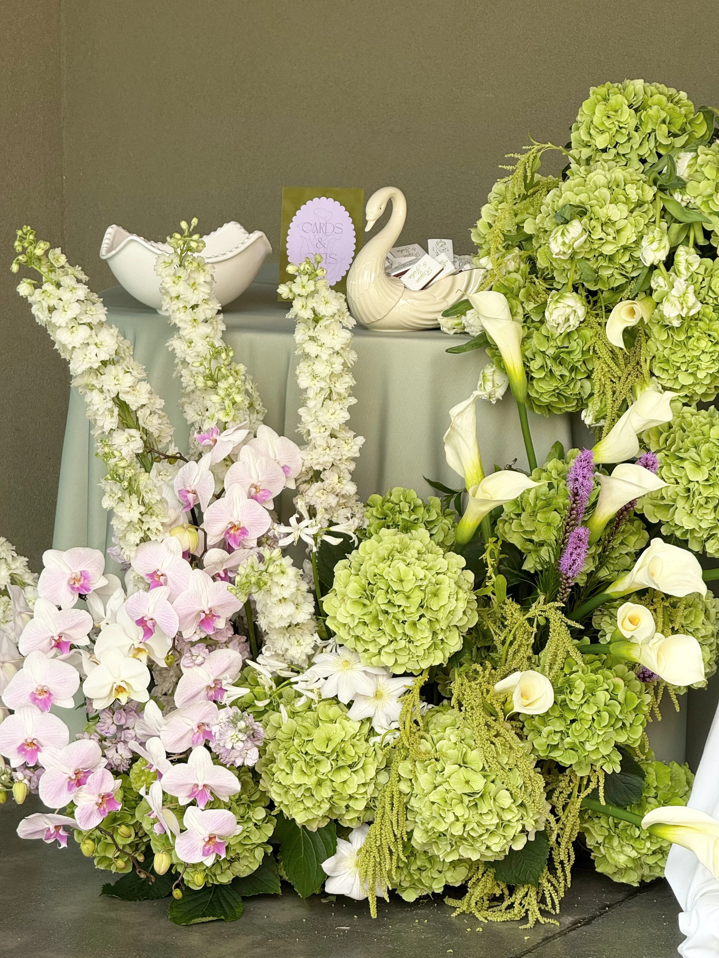 Display of white and green floral arrangements including hydrangeas, calla lilies, orchids, and other flowers on a table with decorative ceramics and a card.