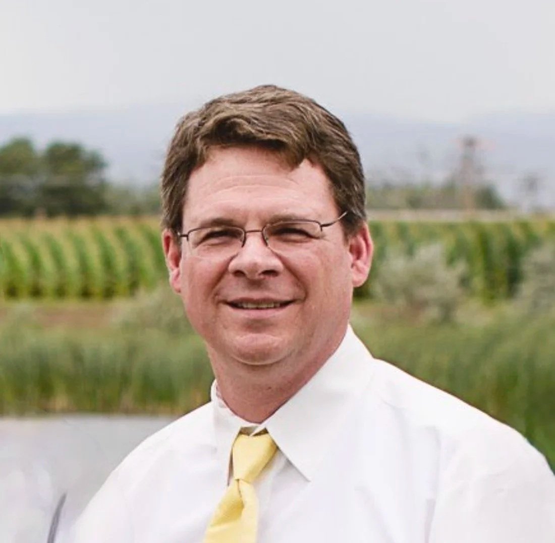 man with brown, hair, glasses, dark suit with white shirt and blue tie, trees in background