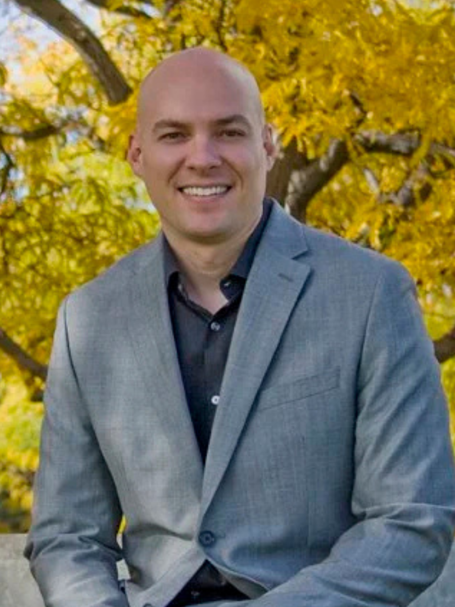 Headshot of bald man, grey suit, black shirt, no tie, yellow tree in background