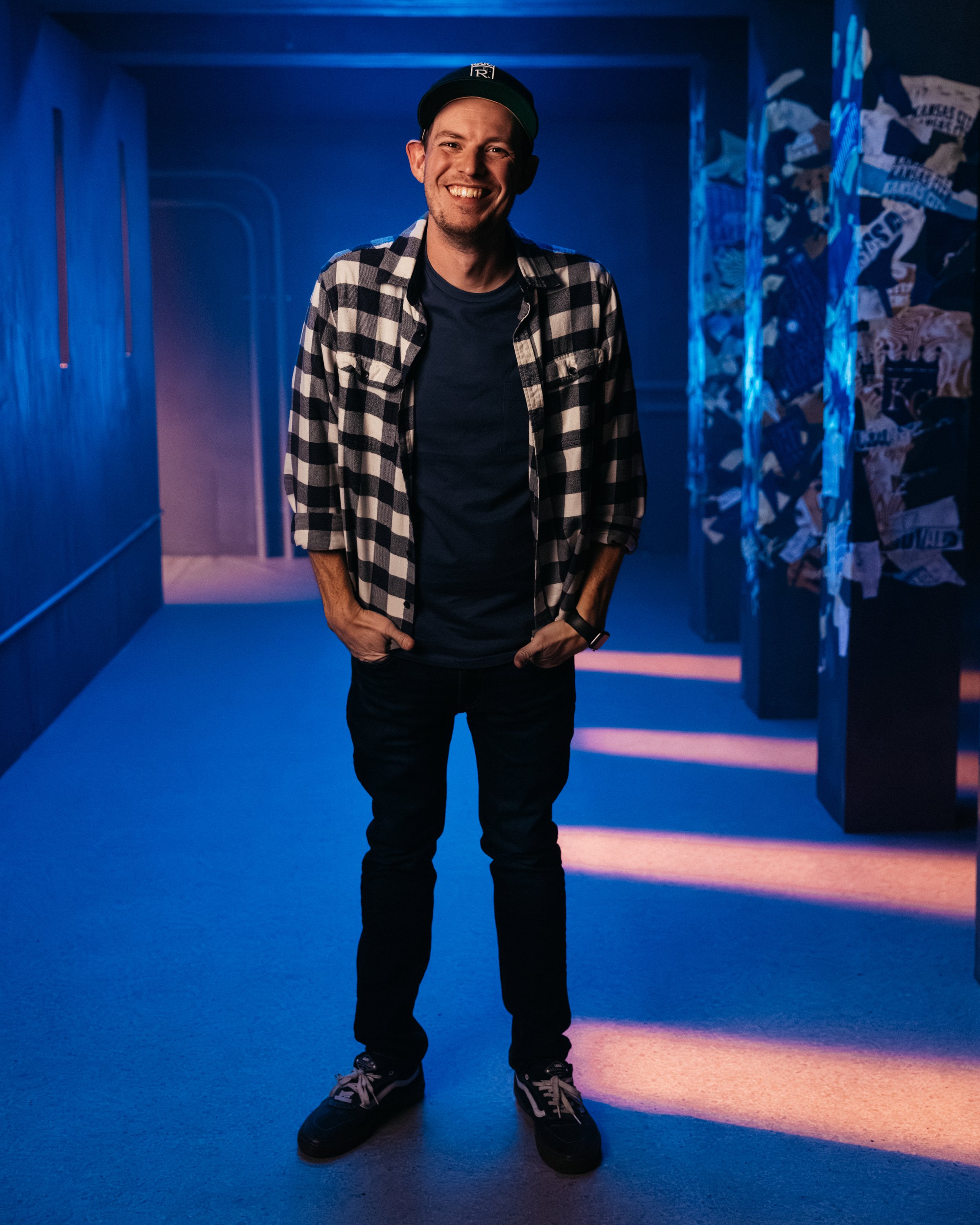 A man smiling and standing in a dimly lit, possibly art installation space with blue and purple lighting. He is wearing a black cap, a black and white checkered shirt, black pants, and sneakers.