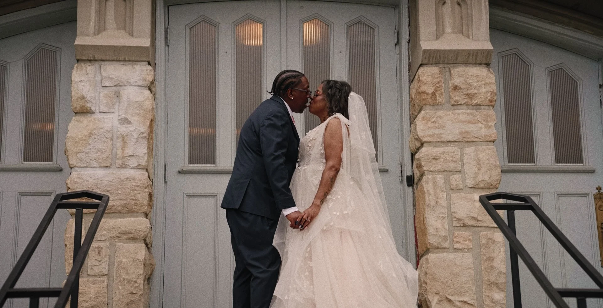A bride and groom sharing a moment outdoors in a cityscape, with the bride holding a bouquet of white and blush roses, and the groom leaning in towards her.