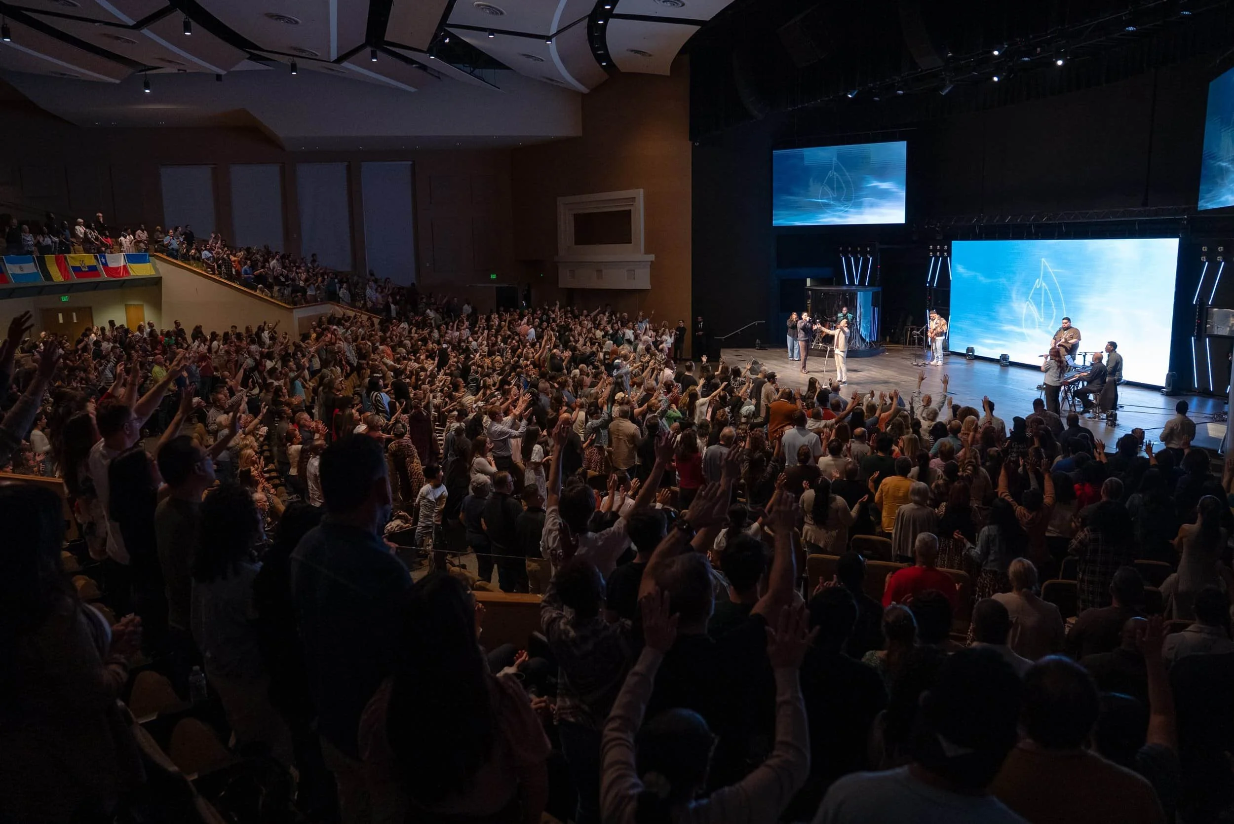 A large crowd with raised hands attends an indoor event with a band performing on stage under blue lighting.