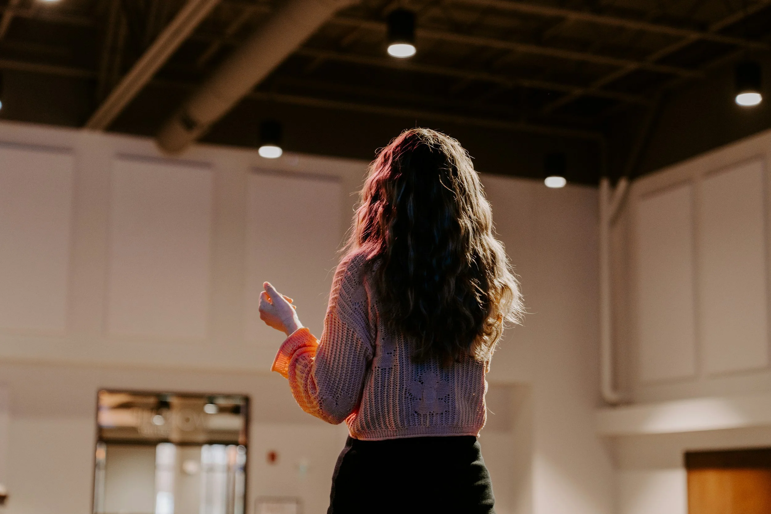 A woman with long, wavy hair wearing a pink knit sweater stands in a large, open indoor space with a high ceiling and overhead lighting. She appears to be gesturing or speaking.