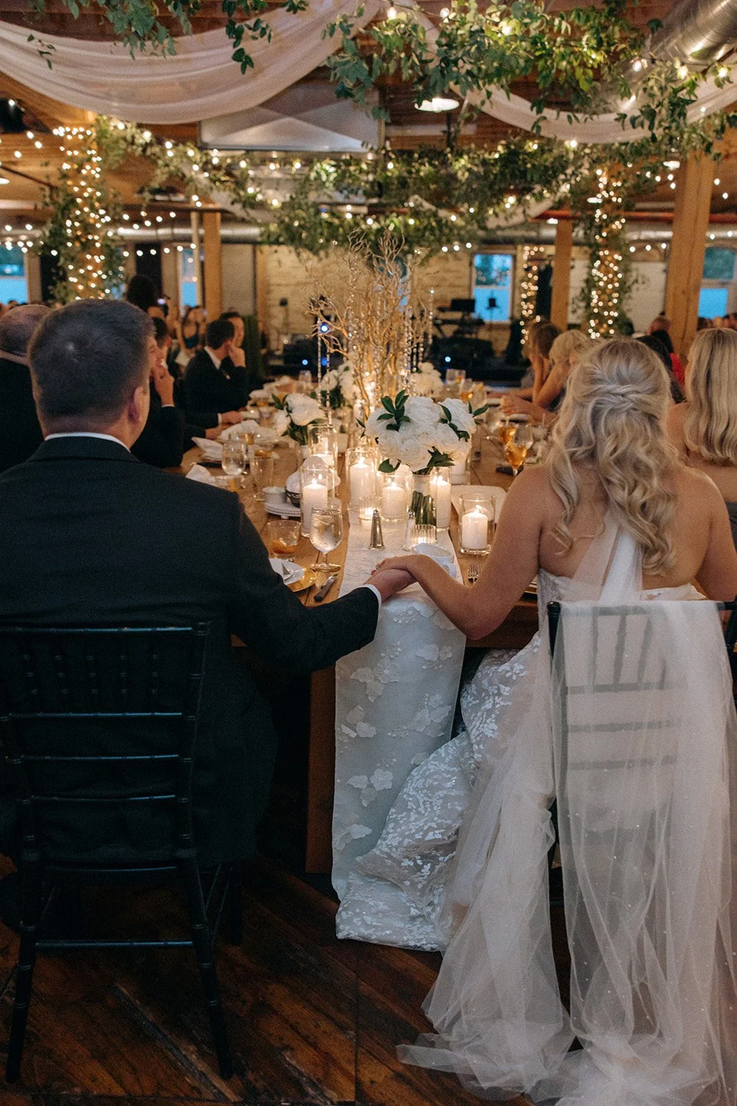 Bride and Groom sit under hanging greenery and draping at wedding reception