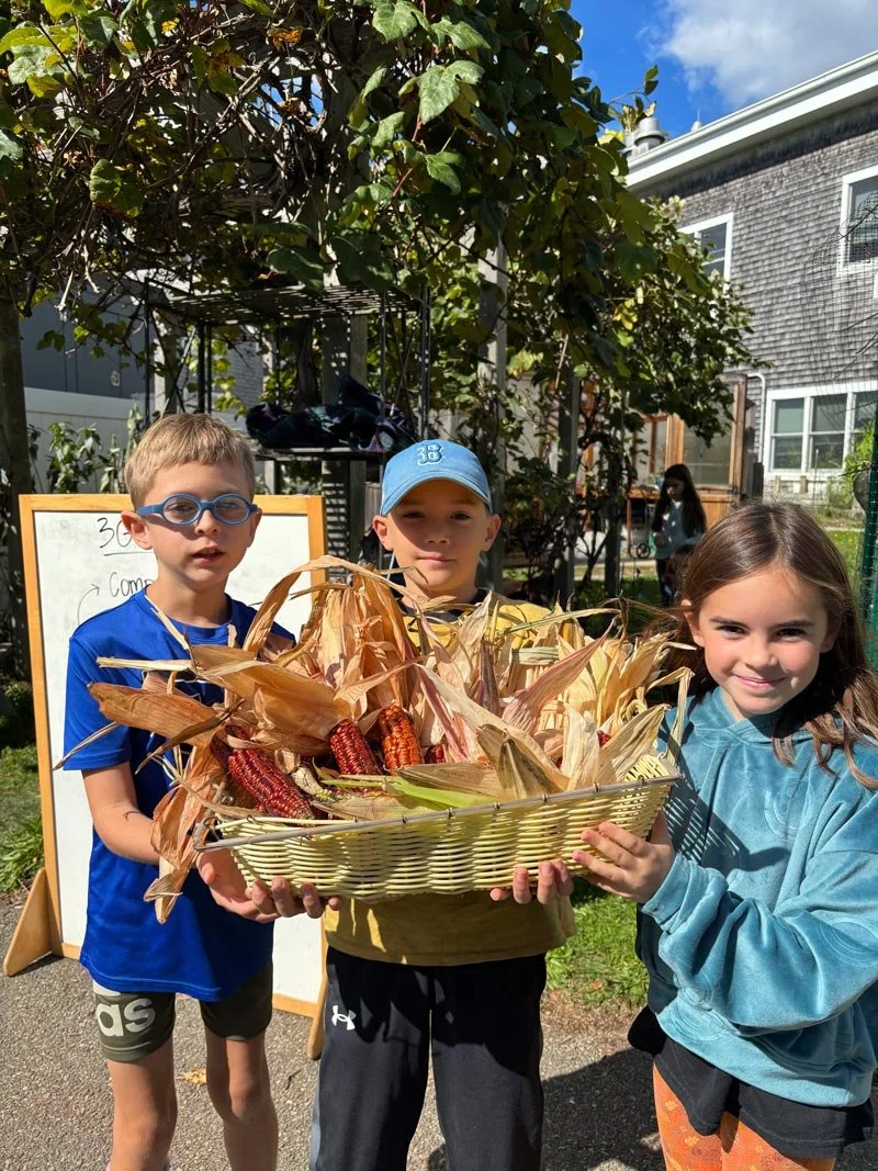 Three children holding a basket of colorful corn cobs outdoors, with a building and tree in the background.