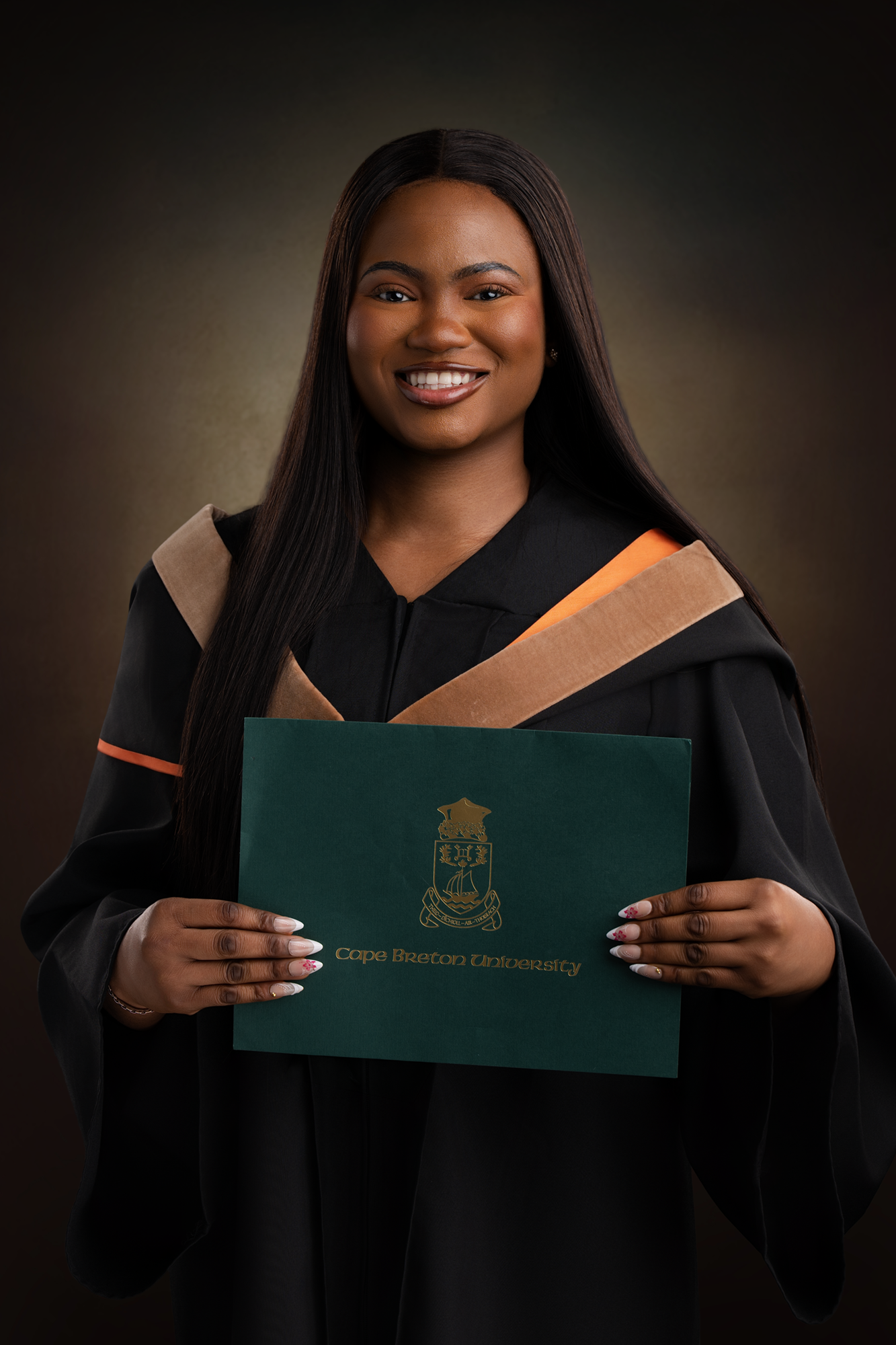 Graduate woman holding Cape Breton University diploma certificate in cap and gown, smiling, with long dark hair and decorated nails, at a graduation ceremony.