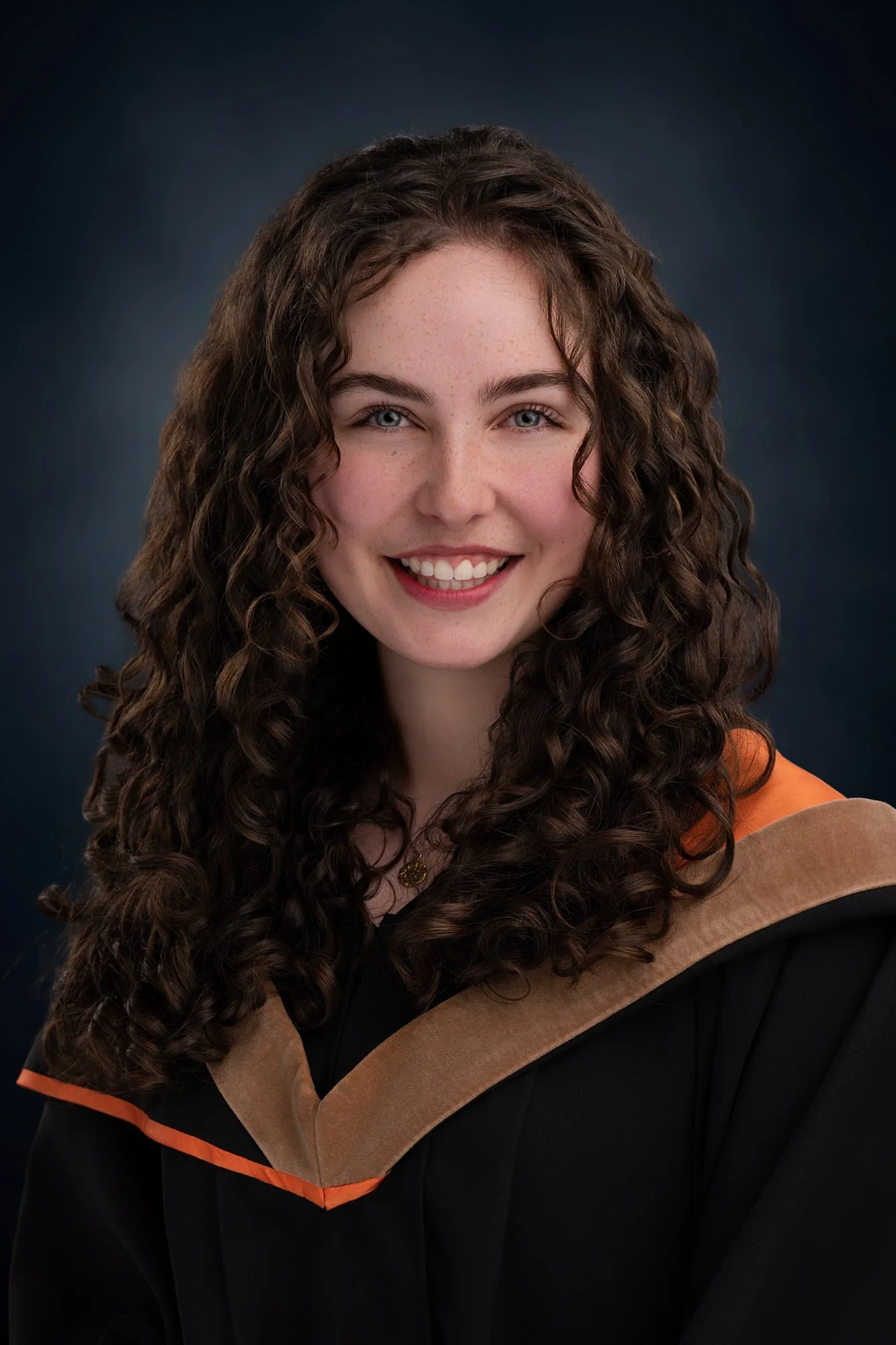 Young woman with long curly brown hair smiling while wearing a graduation gown and hood.