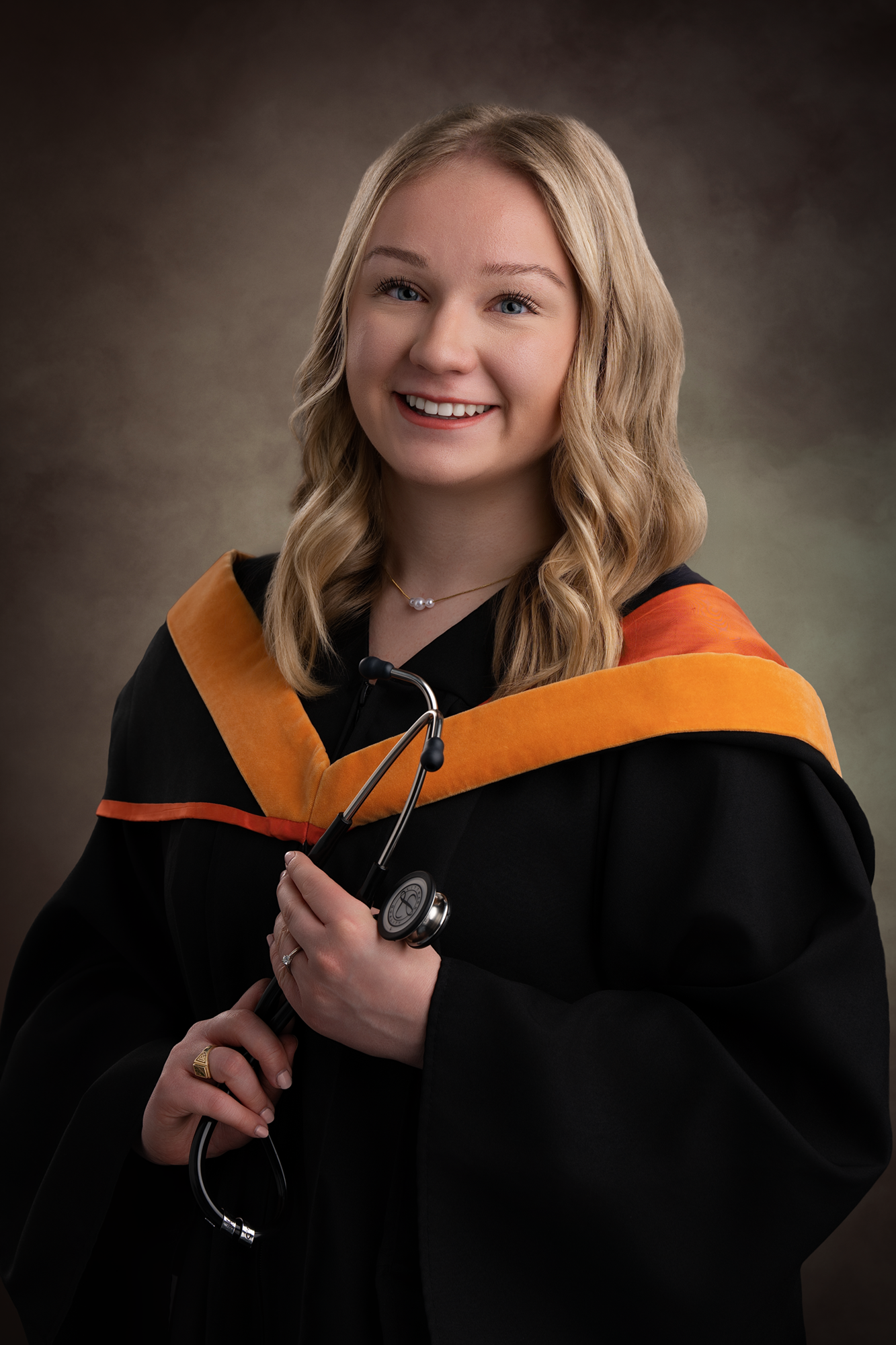 Young woman with blonde hair in a graduation gown with an orange hood, holding a stethoscope, smiling, against a neutral background.