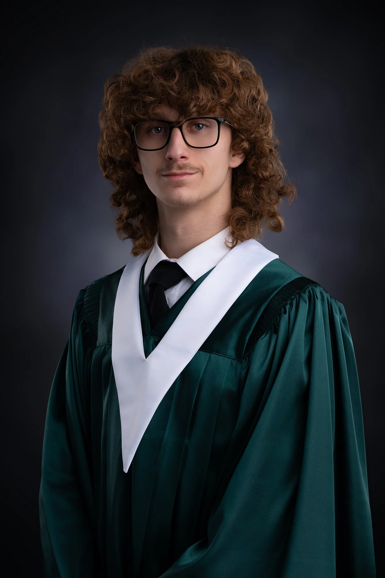 Graduation portrait of a young man from Memorial High School in Sydney Mines, Cape Breton with curly brown hair, glasses, wearing a dark green academic gown with a white and black hood, against a dark background.