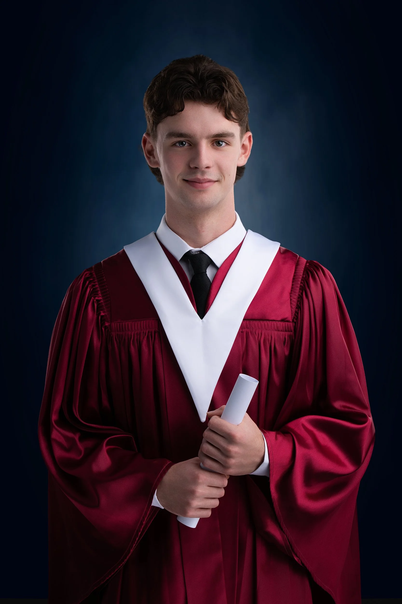 Young man from Riverview High School in Cape Breton in maroon graduation gown with white and black academic hood, holding a rolled diploma, standing against a dark background.
