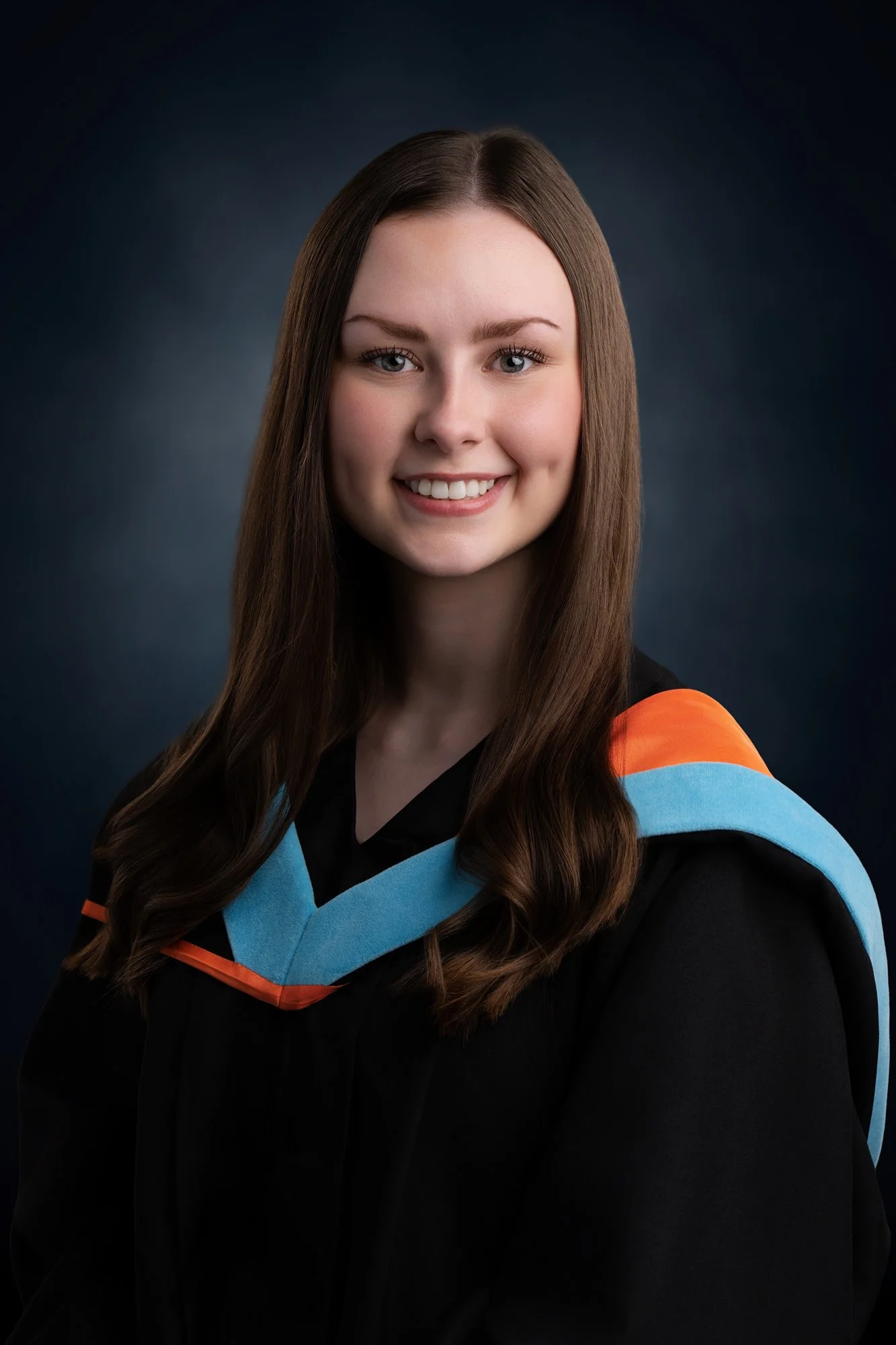 Young woman in graduation gown with blue and orange accents, smiling, against dark background.