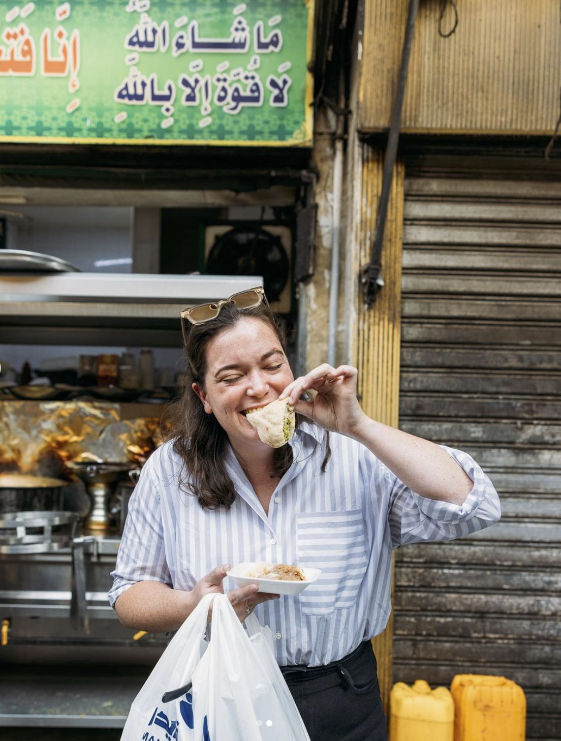 Emily is dressed in a striped button down shirt, standing in front of a food stall in Cairo, Egypt eating a freshly made falafel.